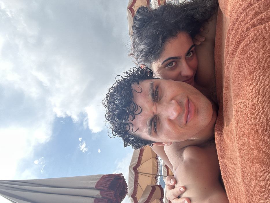 A man and woman with curly hair are lying together on an orange towel, with the woman behind the man. They are both fair-skinned. The man is looking at the camera with a slight smile, while the woman is looking off to the side. There are beach umbrellas in the background, and the sky is visible with some clouds. The photo appears to be taken from above.