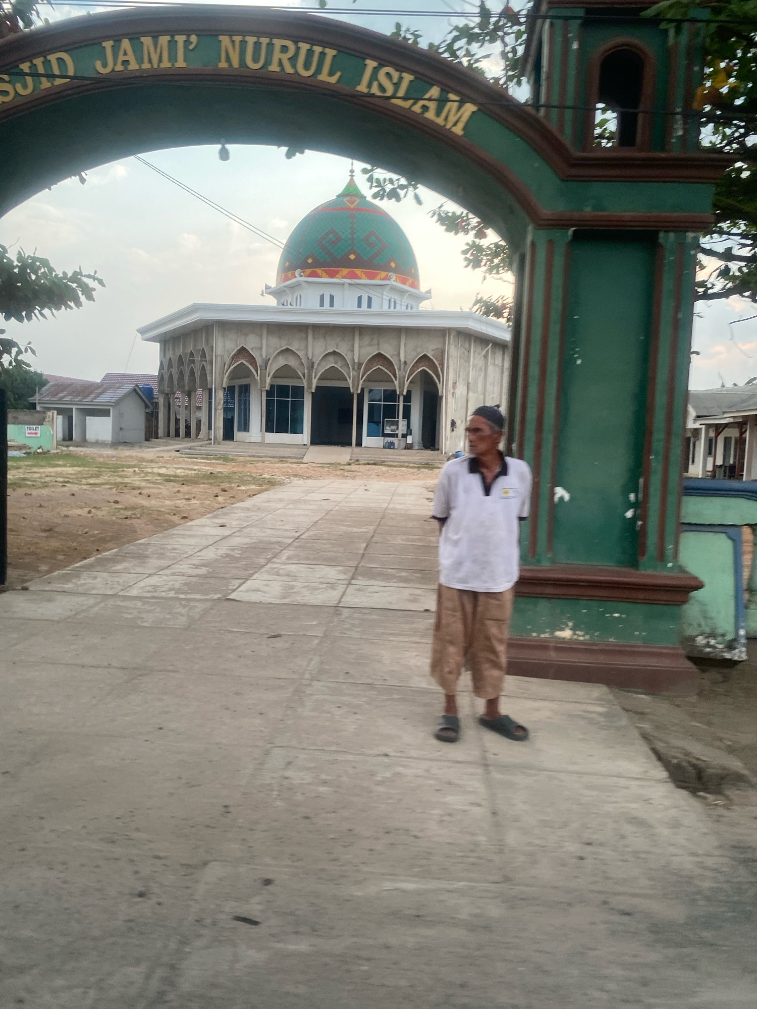 The image captures an exterior view of the Masjid Jami Nurul Islam, located in Banjar Sari, Indonesia. An arched gateway with green and brown trim frames the mosque. The mosque itself is a light-colored building with a prominent green dome, featuring decorative patterns in red and blue. It has a colonnaded facade with arched openings and large glass windows. In front of the mosque, a man stands on a paved pathway. He is wearing a white short-sleeved shirt with a black collar and tan knee-length trousers. He also wears a dark head covering and dark sandals. The ground around the mosque appears to be dirt and gravel, with some vegetation visible. The sky above is overcast.