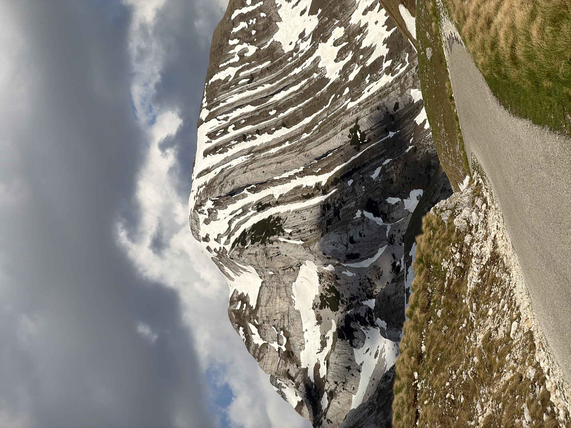 The image presents a view of a rocky mountain partially covered with snow, positioned alongside a narrow paved road. The mountain exhibits distinct horizontal layers, some accentuated with snow deposits. Patches of vegetation are sparsely scattered on the mountain's surface. Adjacent to the road, there's a slope covered with grass and rocks. A portion of the sky, filled with gray clouds, is visible in the background. The location is near the Boričje in Montenegro.