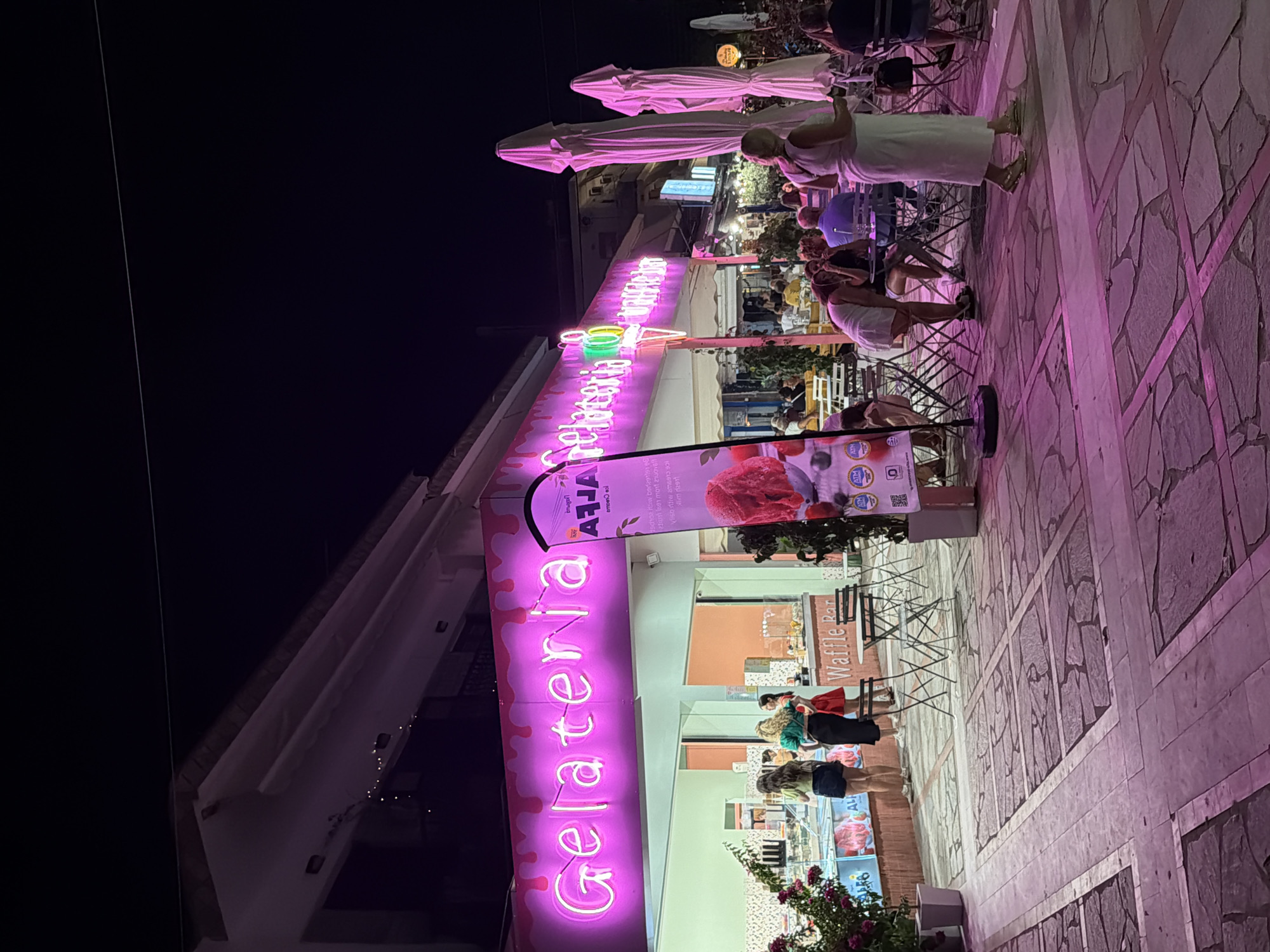 The image depicts a vibrant nighttime scene outside a gelateria and waffle bar in Asprovalta, Greece. A prominent neon sign in bright pink reads "Gelateria" and "Waffle Bar," illuminating the storefront. Several patrons are visible, some queuing inside the gelateria to order, while others are seated at outdoor tables, enjoying their treats. The atmosphere is lively, with people engaging in conversation and enjoying the evening. The architecture of the building is visible, with a white facade and awnings. The ground is paved with large stone tiles, and the scene is further enhanced by decorative lighting and lush green plants in potted containers. The presence of umbrellas suggests the area is designed for outdoor seating.