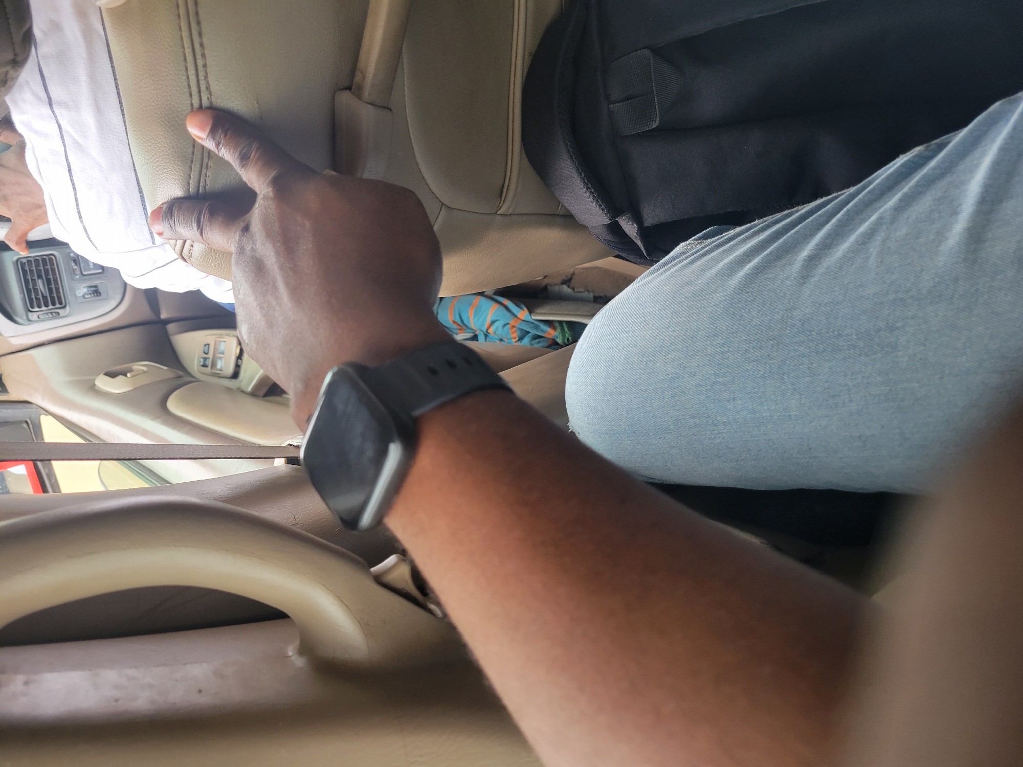 An African person wearing a watch on their left wrist is sitting in a vehicle, possibly a car or bus, in Katambe, Nigeria. Their left arm and hand are visible, with the hand making a peace sign. The person is wearing blue jeans. In the background, another person wearing a white and black striped shirt is visible, sitting in the same vehicle. A black backpack is on the seat behind the person's legs. The interior of the vehicle is visible, including beige upholstery and a car's air conditioning vent. The lighting suggests it is daytime.