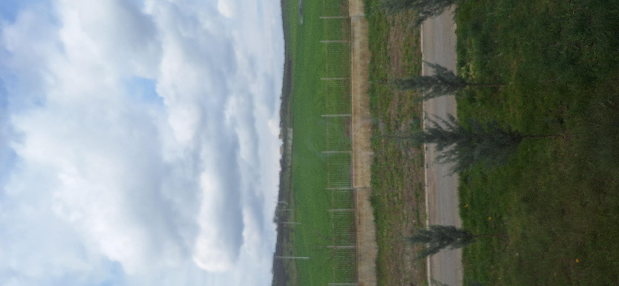 The image shows a landscape scene with a blue and cloudy sky dominating the left side. Adjacent to the sky is a green field with a fence running along its border. Next to the fence is a narrow dirt or gravel path followed by a paved walkway. On the right side of the walkway, there is a grassy area dotted with small trees. The location of this image is Burgas, Bulgaria. There is no evidence to suggest otherwise.