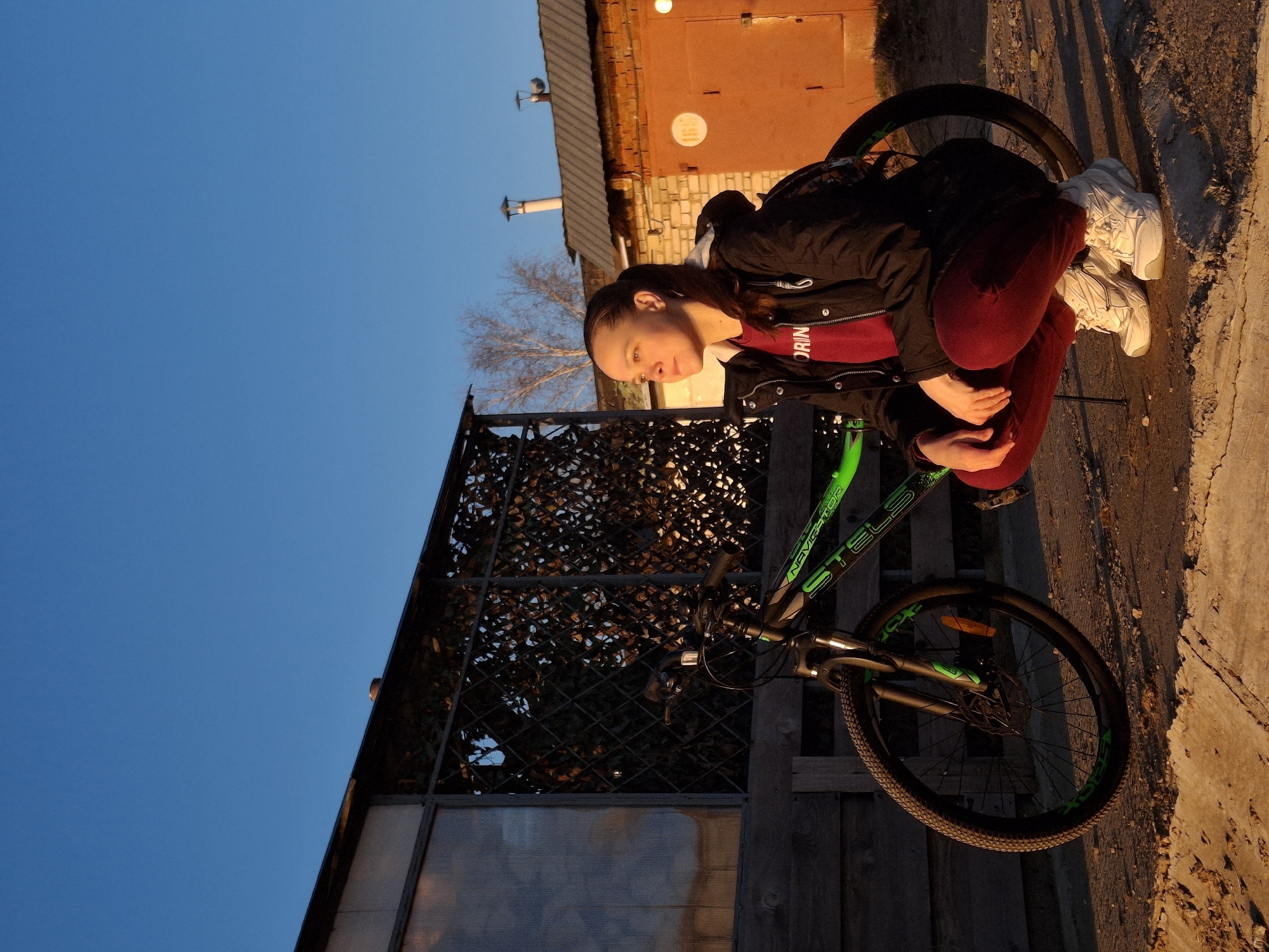 A young woman sits beside a green and black Stels Navigator bicycle, on a sidewalk. The location is near a brick building, possibly a garage, with an orange door and a white circular sign, in Chaikovsky, Russia. The woman wears a black jacket, a red shirt with "ORIN" visible on it, and maroon pants. She is also wearing white sneakers. A building with a metal grill and translucent glass is in the background, next to the bike. The sky is clear and blue, suggesting a late afternoon or early evening setting.