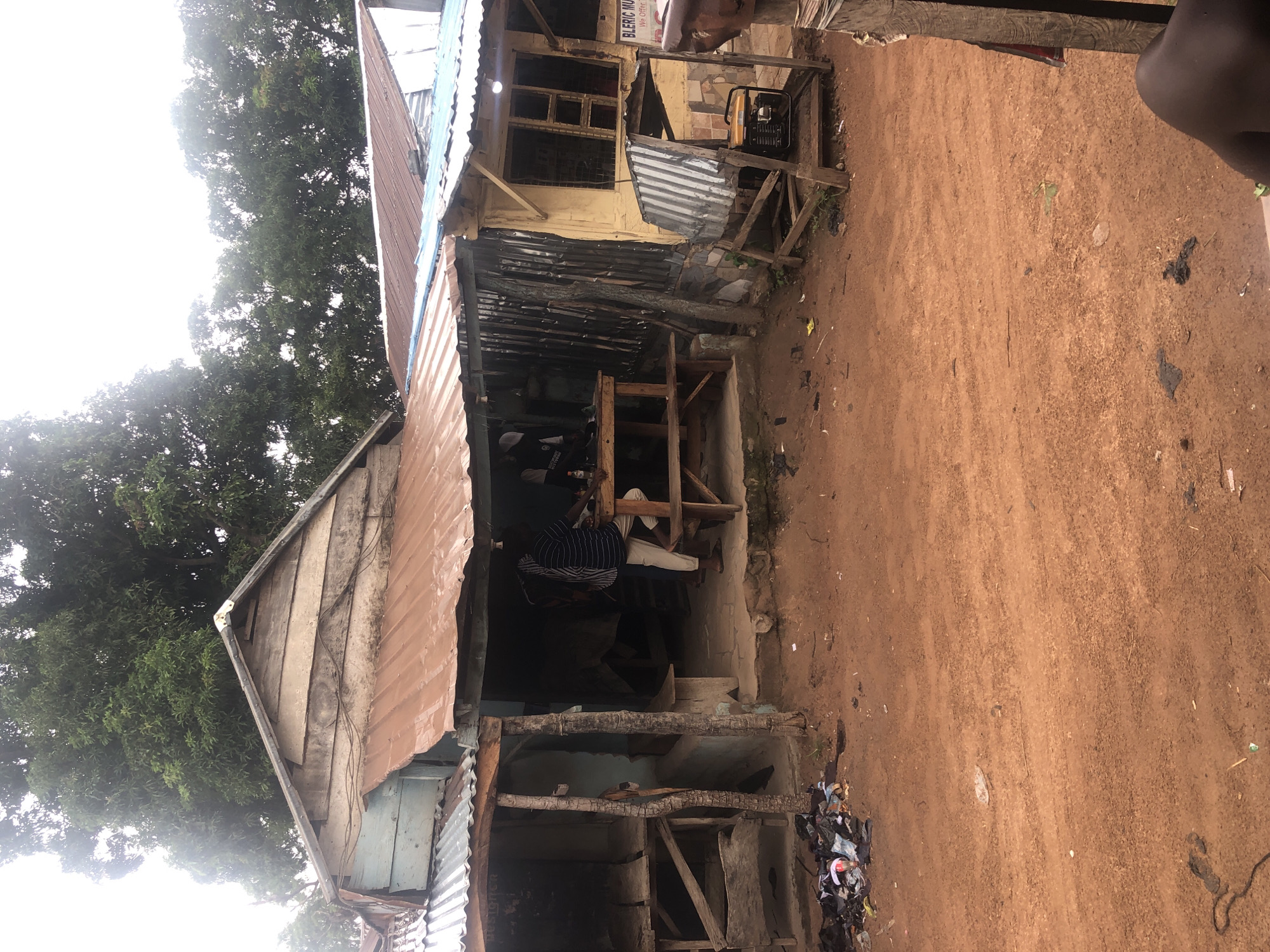 The image depicts an outdoor scene in Wukari, Nigeria. In the foreground, reddish-brown dirt covers the ground, with scattered debris including trash bags and small pieces of refuse. In the middle ground, several makeshift structures with corrugated metal and wooden roofs are visible. One structure has an open front with a wooden table and benches, where two men are present. One man, wearing a striped shirt and light-colored pants, stands near the table. Another man, partially obscured, is also near the table. Adjacent to this structure, another building with a yellow exterior and barred windows is visible. A portable generator is situated under a corrugated metal awning supported by a wooden frame. To the left, a stall with wooden shelves and a partial roof is empty. In the background, lush green trees partially obscure a bright, overcast sky. A sign reading "BLERIC MUSIC" with smaller text below is attached to the yellow building. The overall impression is of a humble, functional marketplace or roadside establishment in an urban or semi-urban environment.