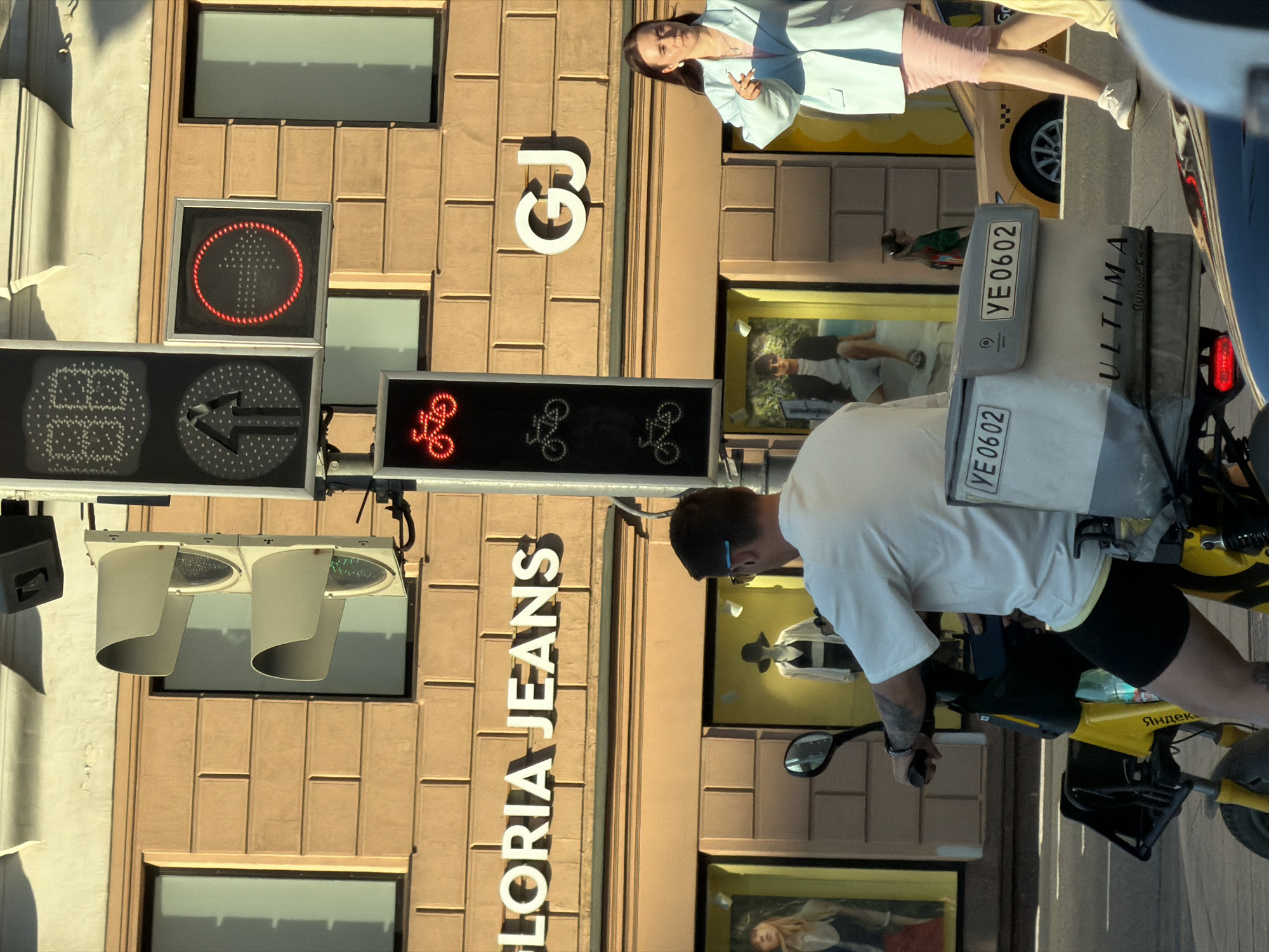 The image shows a street scene in Moscow, Russia, with a building facade in the background. A woman in a light blue jacket and pink skirt stands on a sidewalk, looking towards the camera with a confident expression. In front of her, a person on a yellow electric scooter, wearing a white t-shirt and black shorts, is about to ride away. The scooter has a "Yandex" logo on it, suggesting it is part of a ride-sharing service.
Traffic lights are visible, with one showing a red circle with an arrow, and another displaying red bicycle symbols. The building facade is made of tan brick with large windows. A sign with the words "GLORIA JEANS" is mounted on the building. The time of day appears to be daytime, with sunlight casting shadows. The weather seems to be clear.
Visible text includes "GJ" on the building, "ULTIMA", and the license plate "YE 0602" on a taxi in the background, as well as the "Yandex" logo on the scooter.