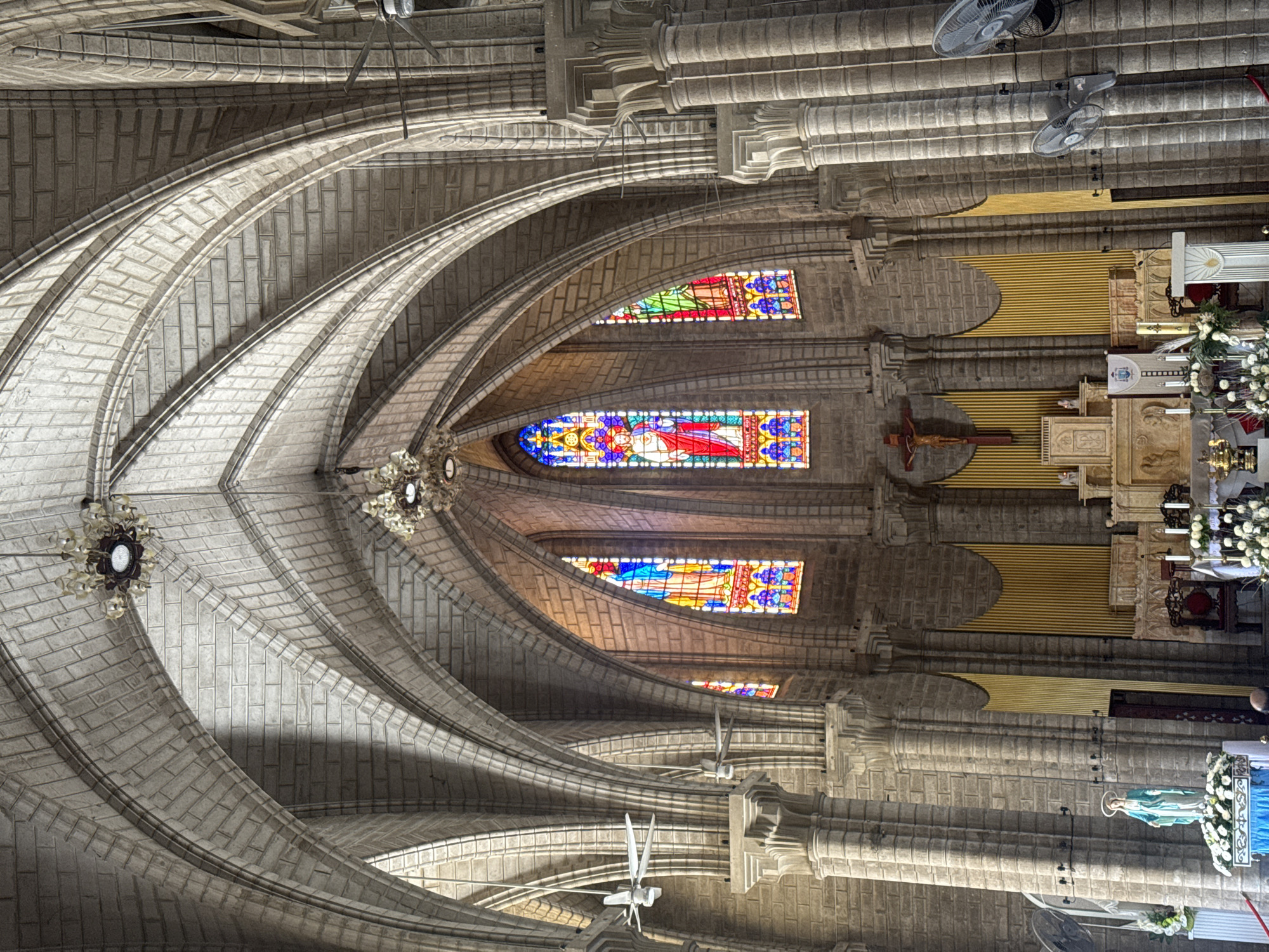 The image depicts the interior of the Nha Trang Cathedral in Nha Trang Ward, Vietnam. The view is directed upwards, showcasing the ribbed vault ceiling constructed from light-colored brick. Stained glass windows are visible along the upper walls, with colors of red, blue, green, and yellow predominating. Architectural elements include columns and arches. A crucifix and a statue of Mary are visible in the sanctuary area. Ceiling fans are mounted on the beams. A chandelier hangs from the center of the vaulted ceiling.