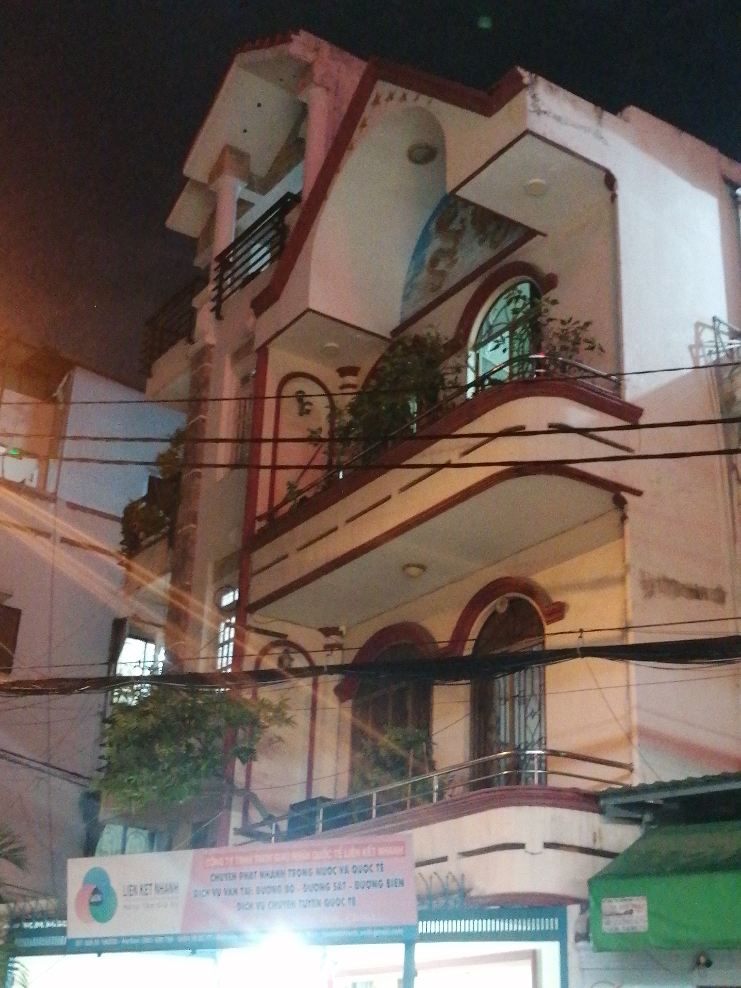The image shows a three-story building at night in Ho Chi Minh City, Vietnam. The building is painted white with red trim and features arched windows and balconies. There are several electrical wires running across the facade. A sign with Vietnamese text and a circular logo is visible on the lower part of the building. The scene appears to be a street in a residential or commercial area.