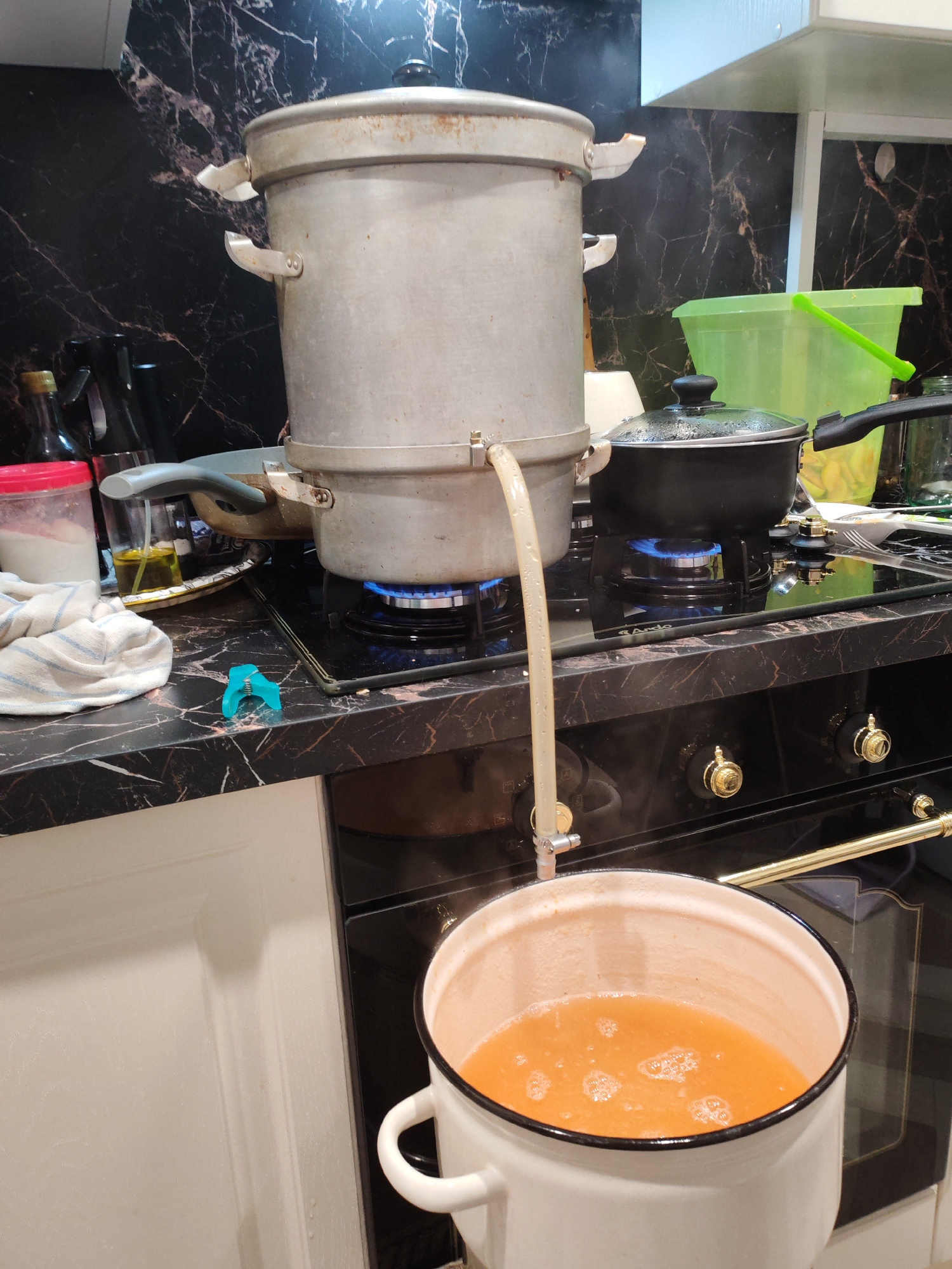 The image shows a kitchen scene with a large metal pot on a stove, connected by a tube to a lower pot containing an orange liquid. The stove is on, with blue flames visible. There are other pots and containers on the counter, including a black pot with a lid and a green bucket. The countertops are black with a marble pattern. The overall setting appears to be a home kitchen. The image is set in the city of Тешевичи, Russia.