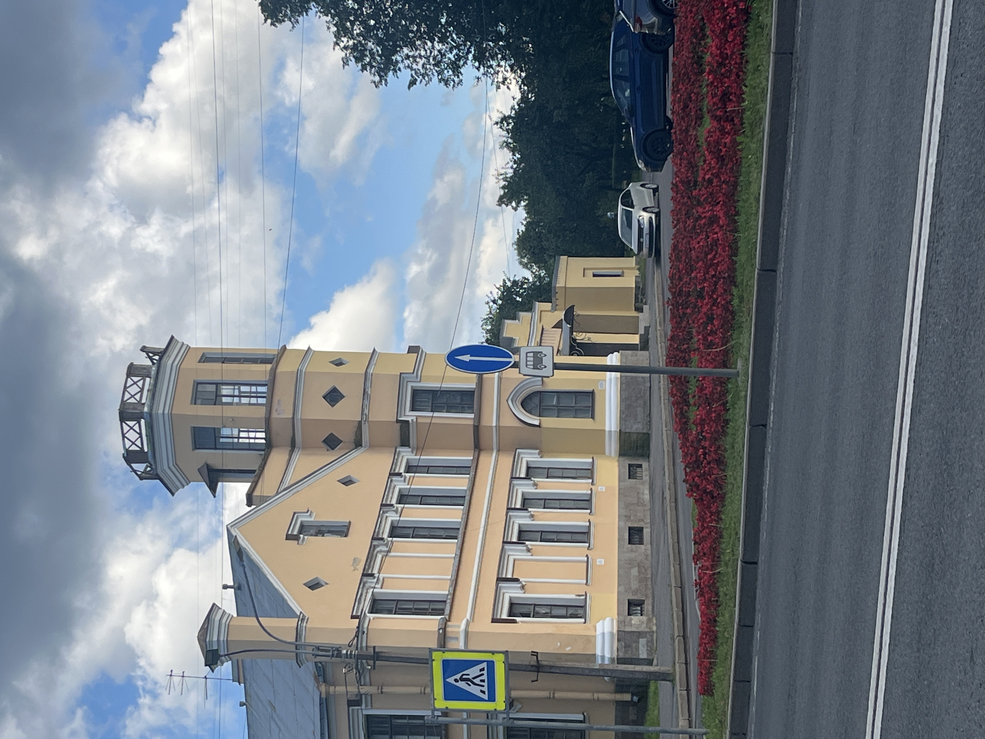 This image depicts a street scene in Peterhof, Russia, dominated by a pale yellow, multi-story building with a distinct tower. The building features numerous windows with white trim and a decorative diamond pattern on its facade. A pedestrian crossing sign is visible on the left, and a traffic sign indicating a bus stop is on the right. In the foreground, a row of vibrant red flowers lines the street, separated from the road by a curb. Several cars are present on the road, including a white sedan and a dark blue SUV. The sky is partly cloudy, with a mix of blue sky and white clouds. The overall environment suggests a bright, daytime scene in a European city.