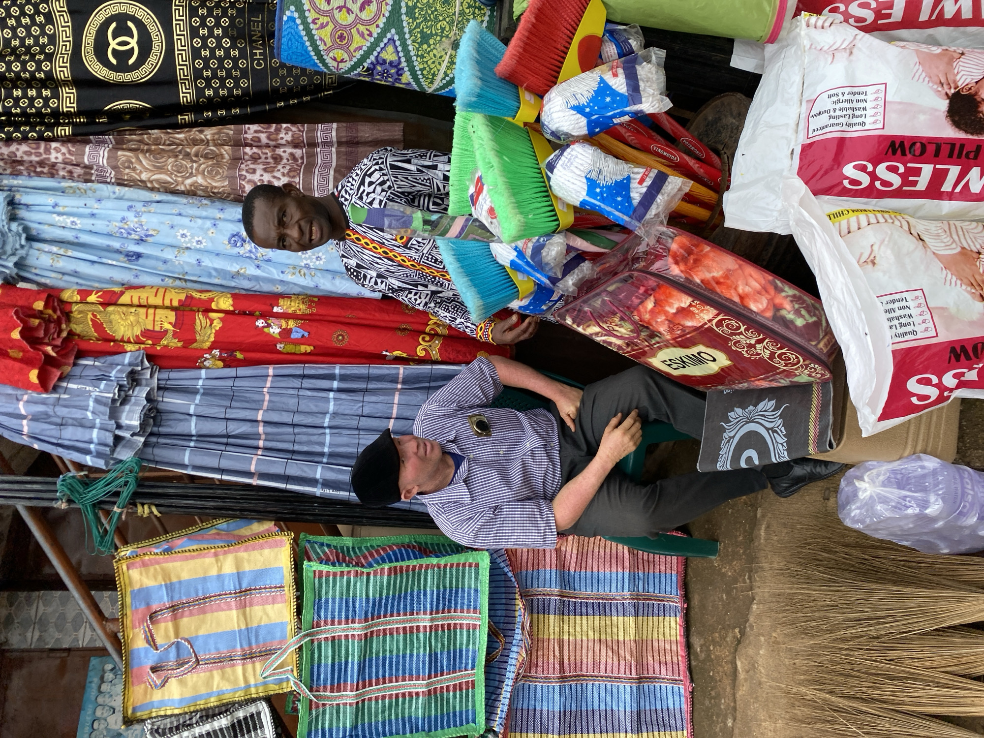 The image depicts a street market stall in Bukuru, Nigeria, showcasing a variety of textiles, household goods, and two men. One man, appearing to be a vendor, is seated on a green plastic chair, wearing a blue and white checkered shirt and dark pants. He has light skin and is wearing a black flat cap. In the background, another man with darker skin, dressed in a traditional patterned top and a colorful necktie, stands in front of a selection of colorful fabrics.
The stall is filled with brightly colored items. Prominently displayed are large, woven striped bags in various color combinations, including yellow, blue, pink, and green. Hanging behind the seated man are draped textiles with intricate patterns, including a black fabric with a gold Chanel logo pattern, a red fabric with a gold dragon motif, and blue fabric with a floral print. In the foreground, a collection of cleaning supplies is arranged, including colorful brooms with blue, green, and red bristles, and packaged mop heads with blue and white packaging. Several large, bagged pillows with pink and white packaging labeled "LESS PILLOW" are also visible. The ground is a dirt surface, and bundles of dried palm fronds, likely for broom making, are stacked on the left side of the image. The overall impression is a vibrant and busy market scene.