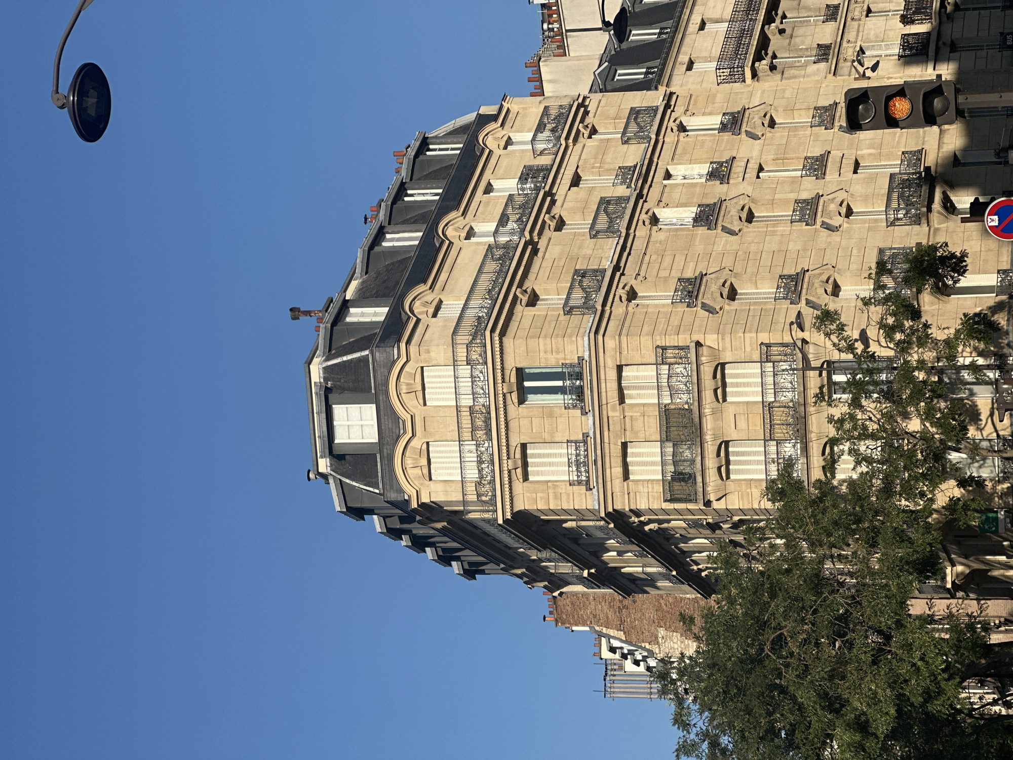 The image captures an upward-angle view of a classic Parisian Haussmannian building under a clear blue sky. The building features a mansard roof with dark grey slate tiles and dormer windows. The facade is constructed from light beige stone, adorned with numerous windows, each framed by white shutters and decorated with ornate black wrought-iron balconies. The building has multiple stories, with balconies on each level.
In the lower right of the frame, a traffic light is visible, showing an illuminated orange light. A street lamp with a dark, circular fixture is positioned above and to the right of the building. To the left, a lush green tree with intricate branches partially obscures the lower section of the building. In the background, other buildings with similar architectural styles, including mansard roofs and chimneys, are visible. The overall scene suggests an urban setting in Paris, France, likely during daylight hours. There are no people visible in the image.