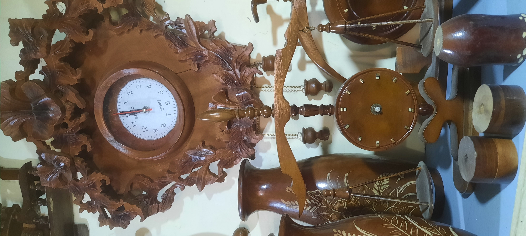 This is a close-up, eye-level shot of a collection of carved wooden objects displayed on a shelf. The most prominent item is an ornate, carved wooden clock with a round face and Roman numerals. The clock is flanked by two decorative carved wooden vases with floral patterns and a decorative wooden balance scale. In the foreground, there are several round wooden objects, possibly for decoration or display. The overall color palette is warm browns and creams, with a slightly muted tone. The lighting appears to be artificial, casting soft shadows. The scene suggests a display of handcrafted items, possibly in a shop or a private collection. The image does not contain any people or specific environmental indicators that would pinpoint its location to Blitar, Indonesia, beyond the provided context.