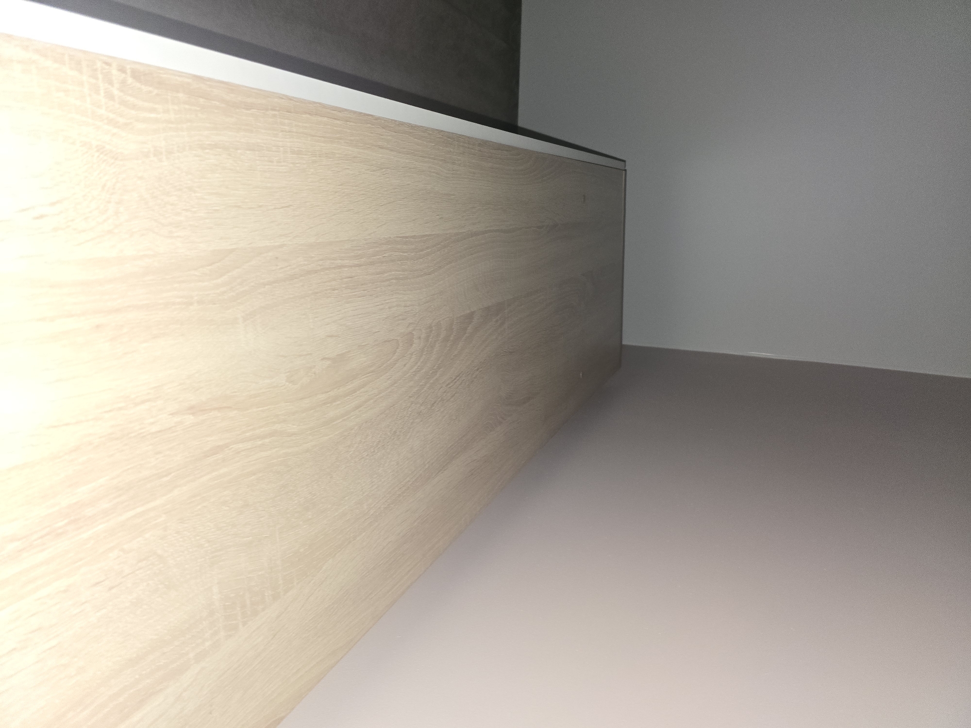 The image shows the top surface of a light-colored wooden countertop, possibly part of a desk or table, with a white edge strip. The countertop is angled, with the foreground wider than the background. To the left of the countertop, a grey floor is visible, and further left, a light grey wall meets the ceiling. To the right and background, a pale pink wall curves away. The scene is indoors, in an interior space. No people or specific identifiable landmarks are present to indicate a location in Ulan-Ude, Russia.