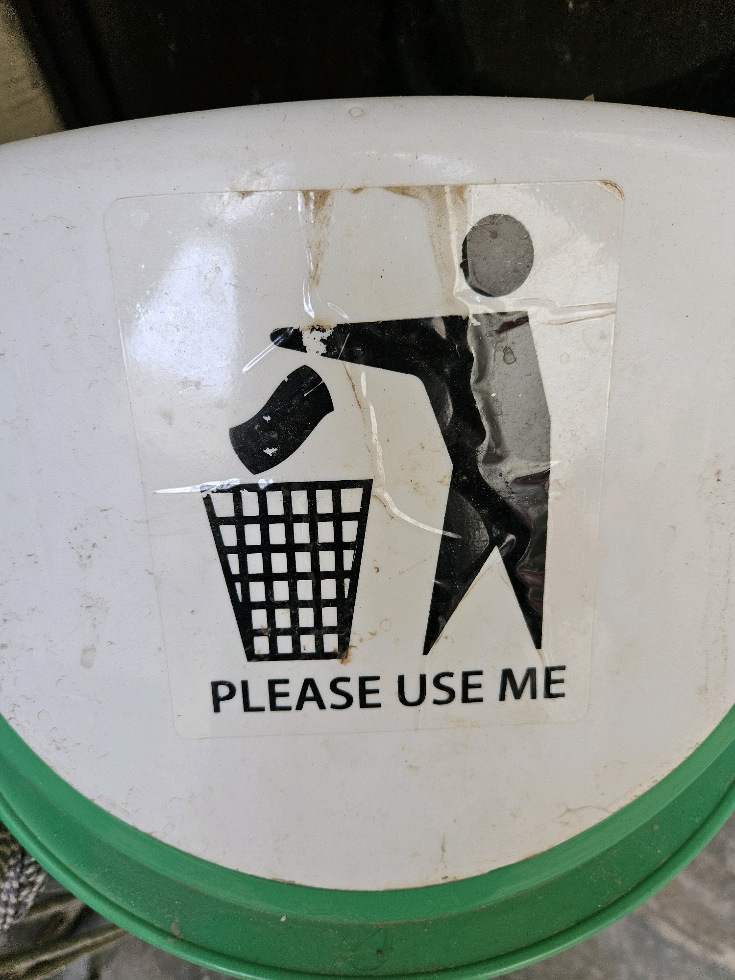 The image displays a close-up, slightly overhead view of a white trash can lid with a green rim. Centered on the lid is a worn, black and white sticker depicting a person throwing trash into a basket. The sticker has been partially covered by a clear protective film which is peeling and creased in places, and there are visible smudges of dirt and grime on both the sticker and the lid. Below the graphic, the text "PLEASE USE ME" is printed in black capital letters. The background is dark and out of focus, providing no discernible context about the surrounding environment or its location within Jawad Avenue, Pakistan.