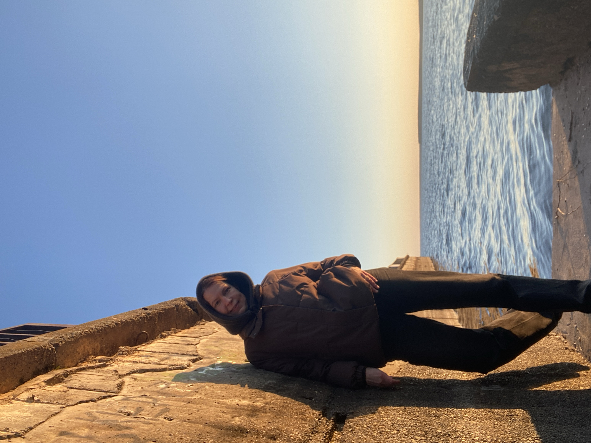 A photograph captures a woman lying on a concrete structure adjacent to water, possibly a river or sea. She is wearing a dark brown puffer jacket with the hood up, along with dark pants and black shoes. The woman is smiling slightly and appears relaxed. The concrete structure she is on is uneven and appears to be a pier or embankment. The water is visible in the background, with a strip of lighter, almost golden sky above it. The upper left corner of the image shows a clear blue sky. The photograph was taken in the city of Chaikovsky, Russia.