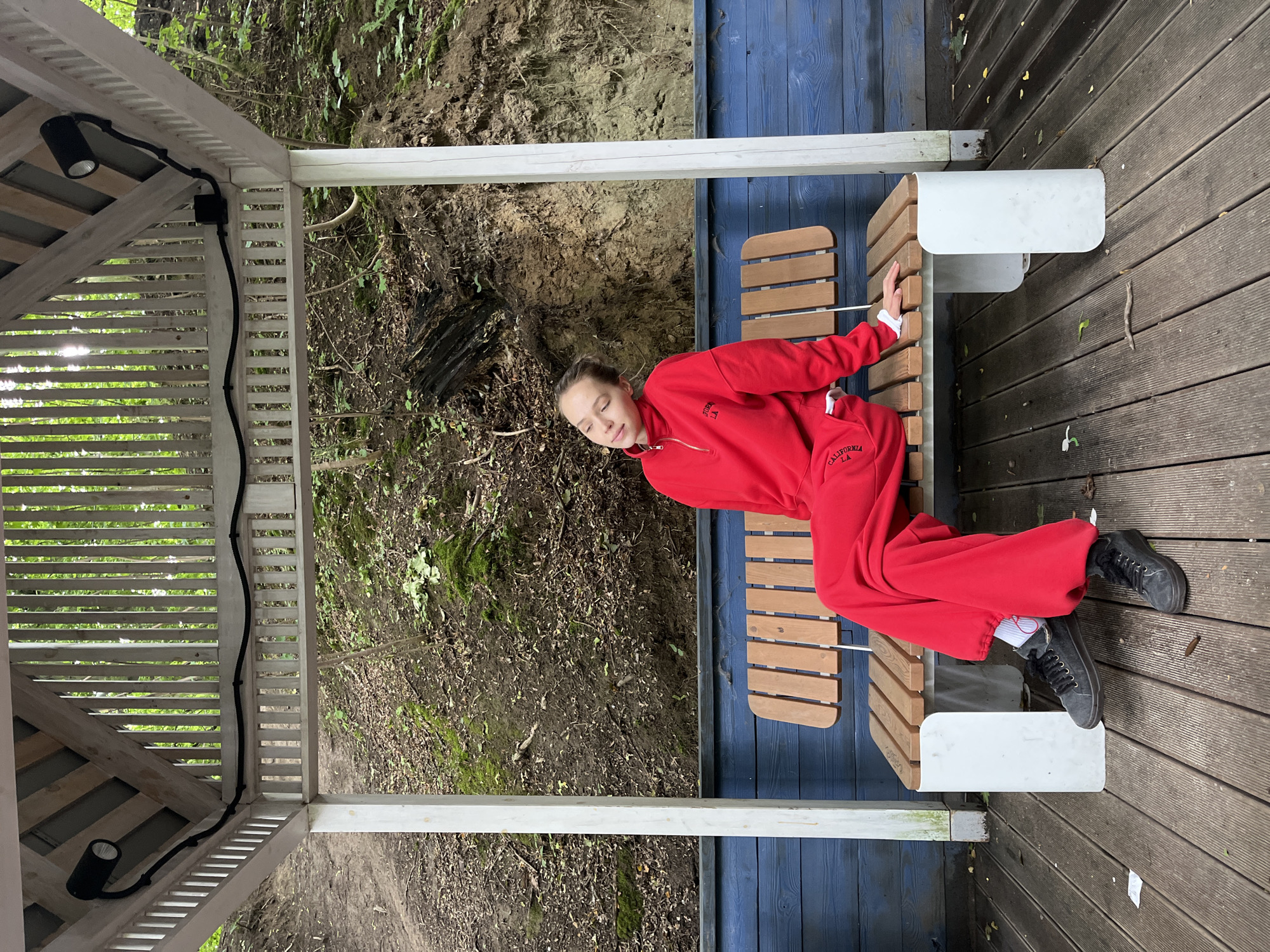 A young woman wearing a red tracksuit and black sneakers sits on a wooden bench in a gazebo. She has her eyes closed and her head tilted back. The gazebo has a wooden roof and a blue wooden wall behind the bench. The ground is covered with wooden planks. In the background, there is a wooded slope with moss and fallen leaves. The scene appears to be in a park or natural area. The woman is sitting alone on the bench.