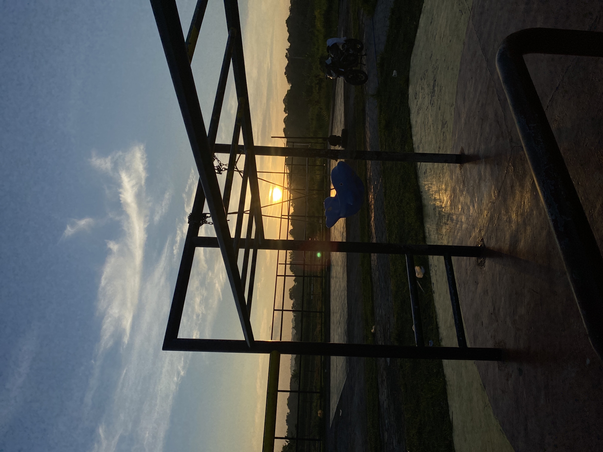 A playground merry-go-round structure stands in silhouette against a sunset sky, located in Davao City, Philippines. The sun is setting behind a line of trees in the distance, casting a warm glow on the scene. A blue, whale-shaped swing hangs from the structure, and a motorcycle is parked to the right of the frame. The ground around the merry-go-round is paved with concrete and grass. The sky is a mix of blue and orange hues, with wispy white clouds. The scene appears to be a public park or recreational area.
