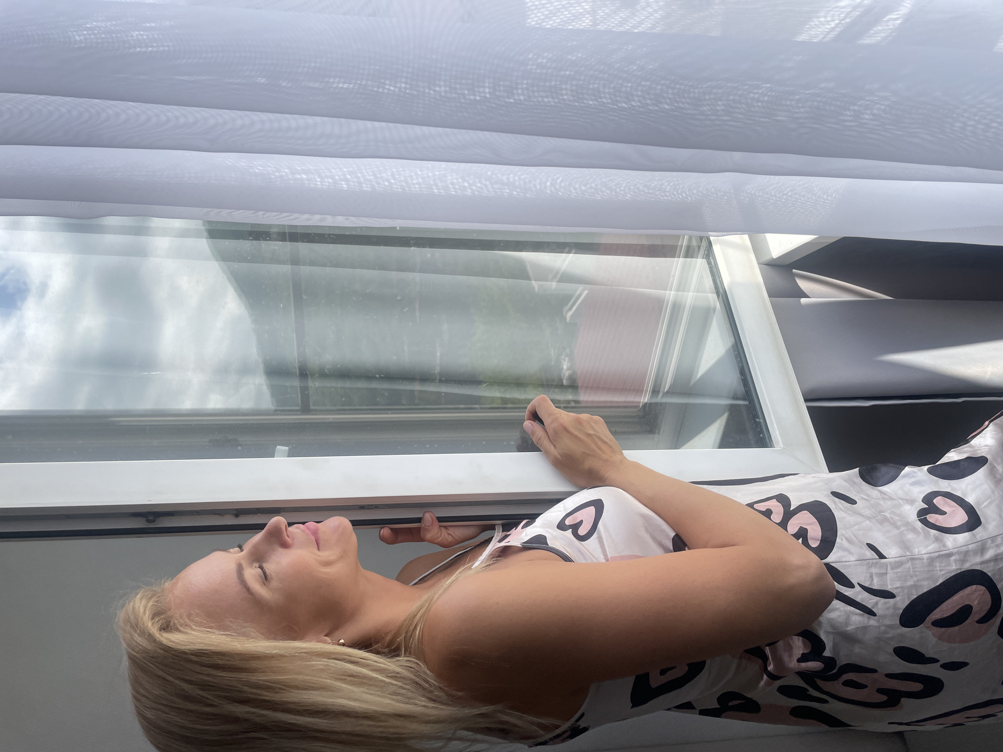 A woman with blonde hair is lying down next to a window, looking upwards. She is wearing a white and pink heart-patterned top. The window has a white frame, and translucent white curtains are visible above it. The background visible through the window shows an outdoor scene with a cloudy sky. The scene does not provide any details to indicate it is in the city of Baranovka, Russia.