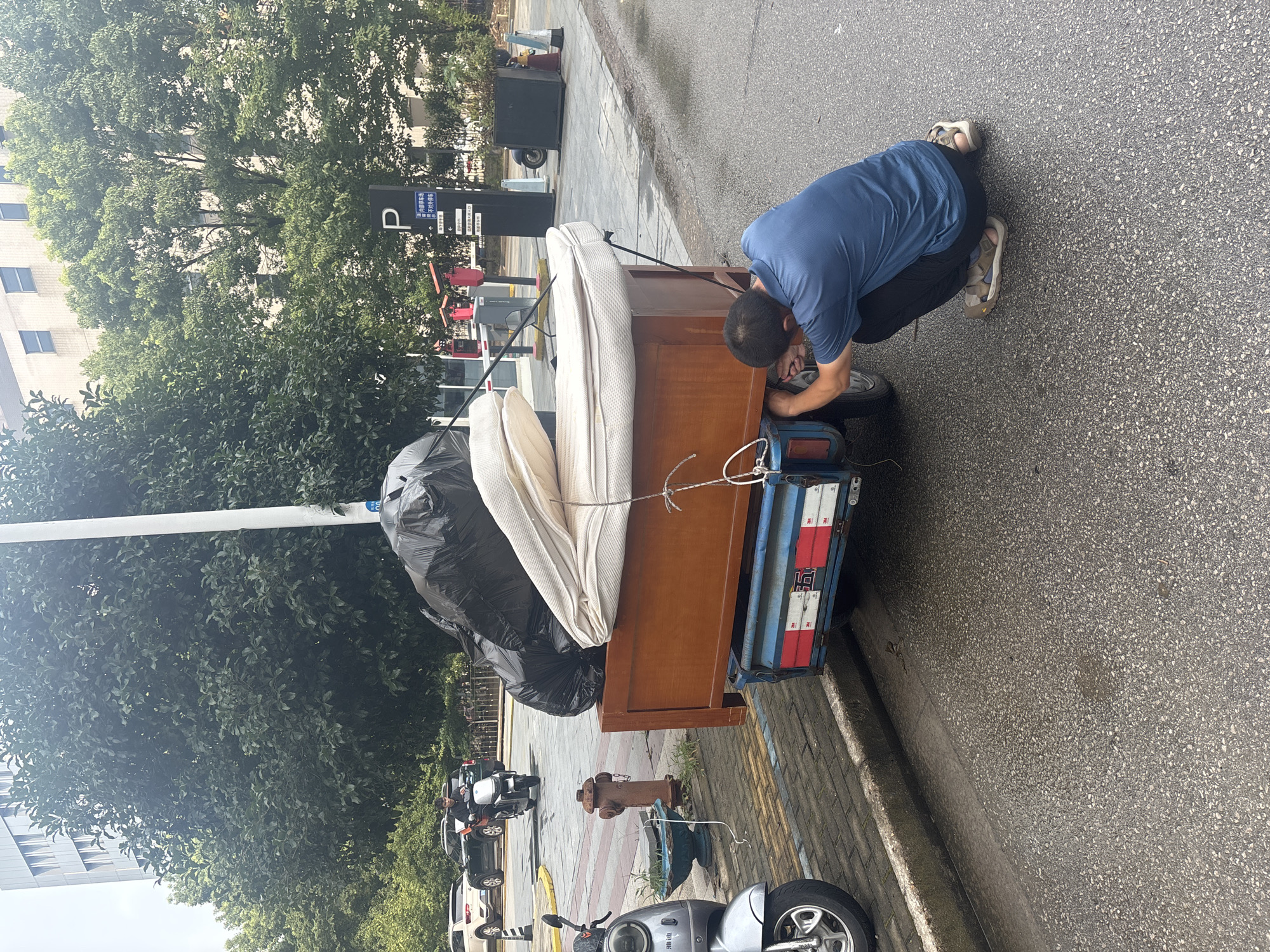 In Suzhou Industrial Park, China, a man wearing a blue t-shirt and dark pants is squatting by the back of a three-wheeled vehicle. The vehicle is loaded with household items, including a large black trash bag, a mattress, and what appears to be a wooden headboard. The man is actively working on the rear wheel of the vehicle, suggesting a mechanical issue. The scene takes place on a paved road next to a sidewalk and a grassy area with trees. In the background, there are parked cars, a motorcycle, a fire hydrant, and a parking structure with a "P" sign. The sky is overcast, and the ground is wet, indicating recent rain.