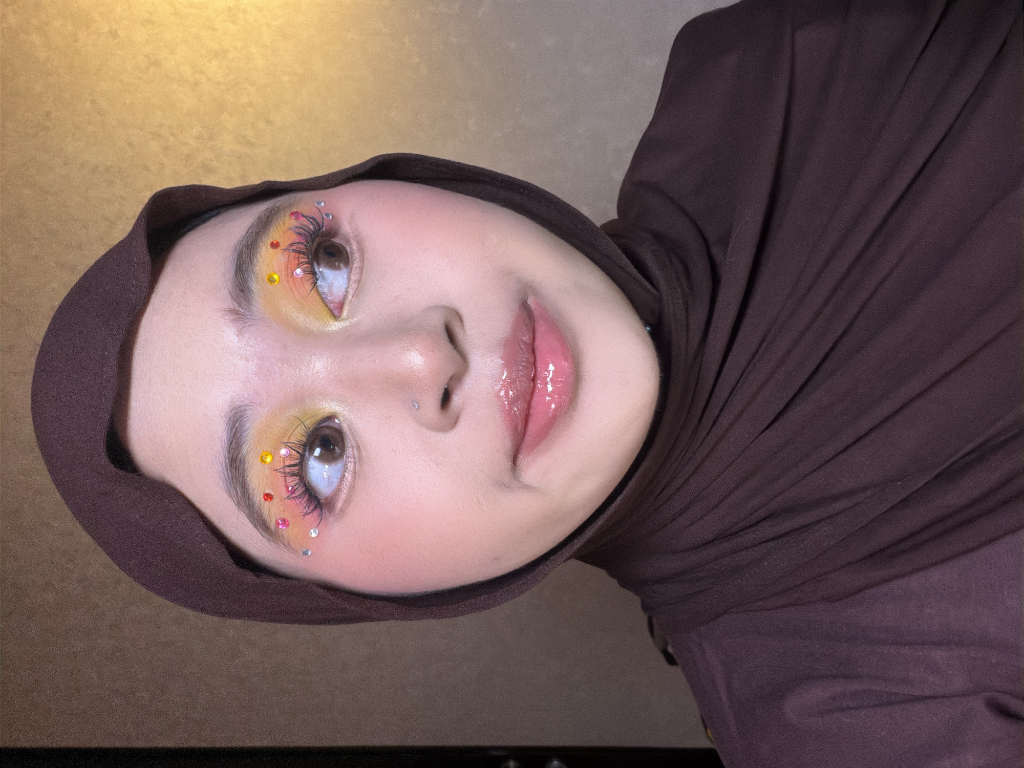 A medium shot shows a young woman wearing a brown hijab and a brown draped top. She has on dramatic eye makeup with yellow and pink eyeshadow, false eyelashes, and colorful rhinestones applied around her eyes. Her lips are glossy and pink, and she has a small nose piercing. She is looking off to the right side of the frame with a slight smile. The background is a textured wall with a gradient of warm tones, transitioning from a darker brown on the left to a lighter, golden hue on the right. The lighting is soft, highlighting her facial features and makeup.