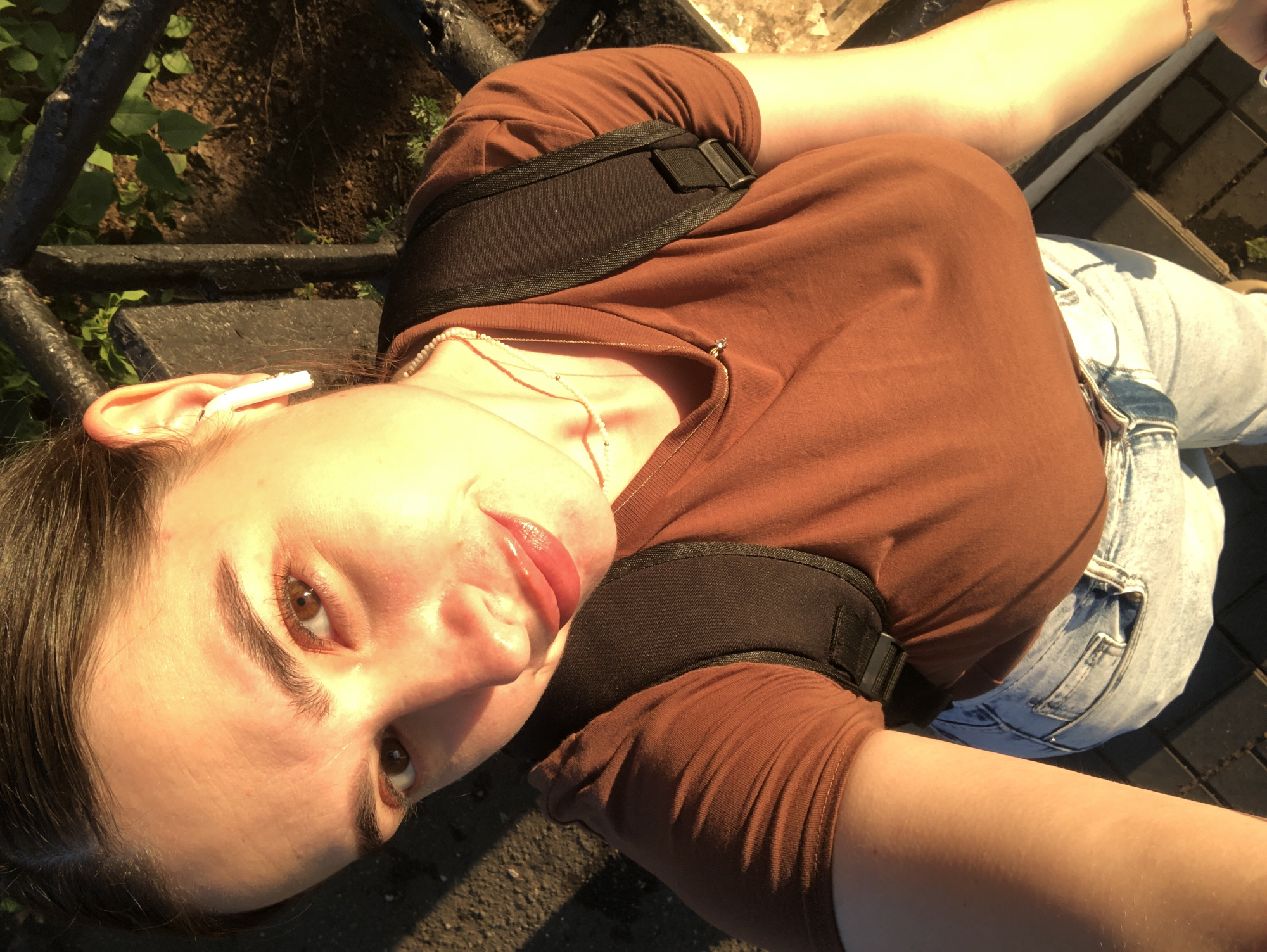 A young woman, facing the camera, stands outdoors in Chaikovsky, Russia, in what appears to be a park or public space, illuminated by golden hour sunlight. She has dark hair pulled back and is wearing a brown t-shirt, light blue jeans, and a black backpack. A white wireless earbud is visible in her right ear. She is also wearing two delicate necklaces. The background shows a dark metal railing and greenery. The ground beneath her is paved with brick-like stones.