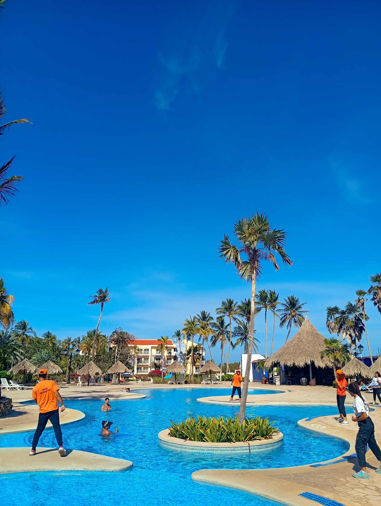 The image depicts a bright, sunny day at what appears to be a resort pool in Playa El Agua, Venezuela. The sky is a vibrant blue with scattered clouds. A large, multi-sectioned pool with varying depths occupies the foreground and middle ground. Small islands with palm trees and foliage dot the center of the pool. Several people are either in the pool swimming or standing on the surrounding paved areas.
Two individuals wearing orange shirts with "TEAM ANIMACIÓN" printed on them are positioned near the pool, possibly as resort staff or activity instructors. One is walking towards the pool and the other is standing near a palm tree. Additional people can be seen swimming in the pool and standing or walking near the pool's edge.
In the background, a multi-story building with a terracotta-colored roof is visible behind the palm trees and thatched-roof structures. These structures likely serve as poolside bars or lounging areas. Palm trees are abundant throughout the scene, contributing to the tropical ambiance.