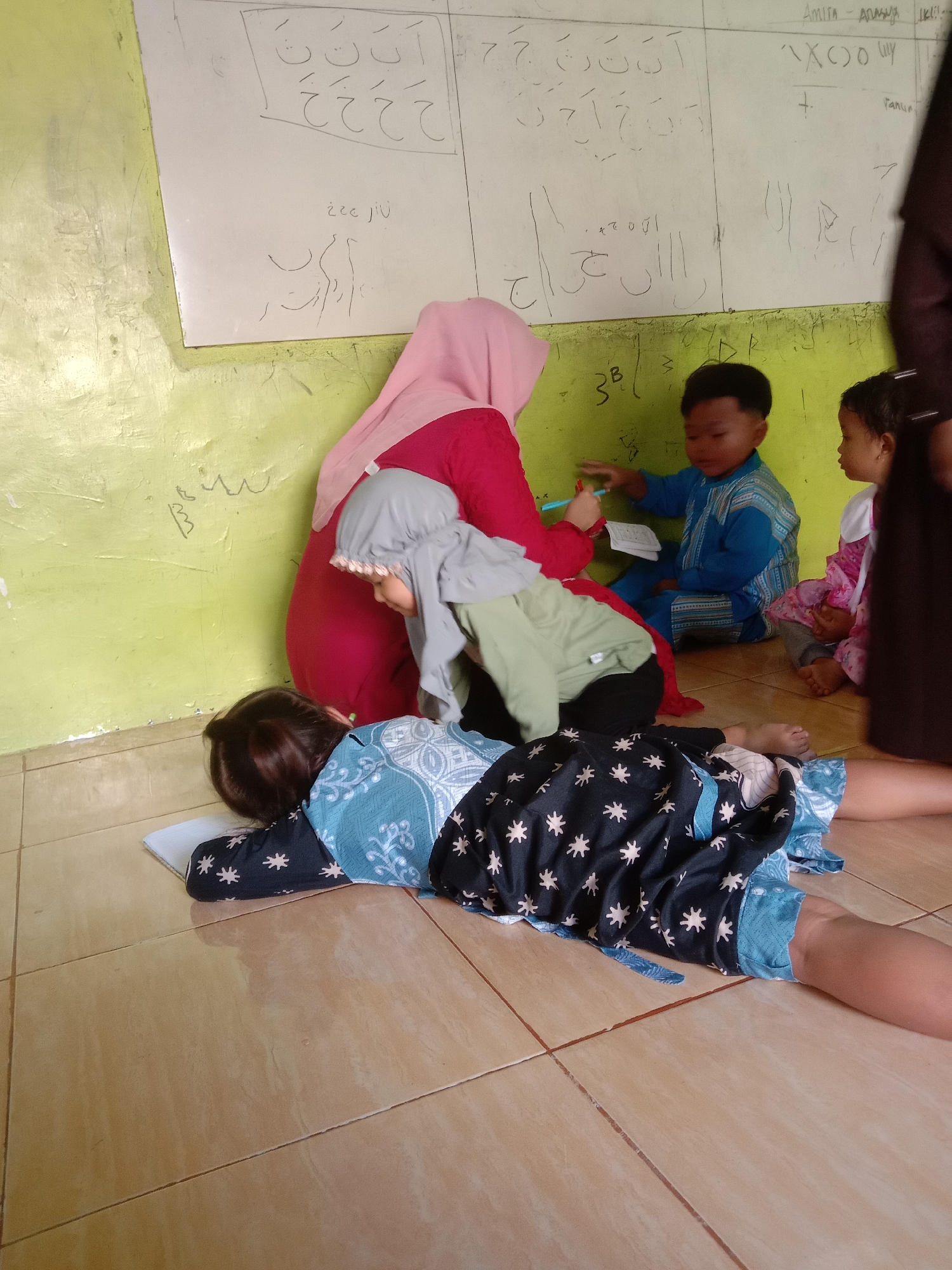 In Tulungrejo, Indonesia, a group of young children and an adult are gathered around a whiteboard, presumably in a classroom. The adult, wearing a pink hijab and red top, is interacting with a boy in a blue shirt who is sitting on the floor and pointing at the whiteboard. Another child in a grey hijab is looking towards the adult. In the foreground, a child lies on their stomach on the floor, facing away from the camera, with their head resting on a white mat or book. The child is wearing a patterned black and blue outfit. The room has green walls and tiled floors. The whiteboard contains handwritten Arabic script and numbers.