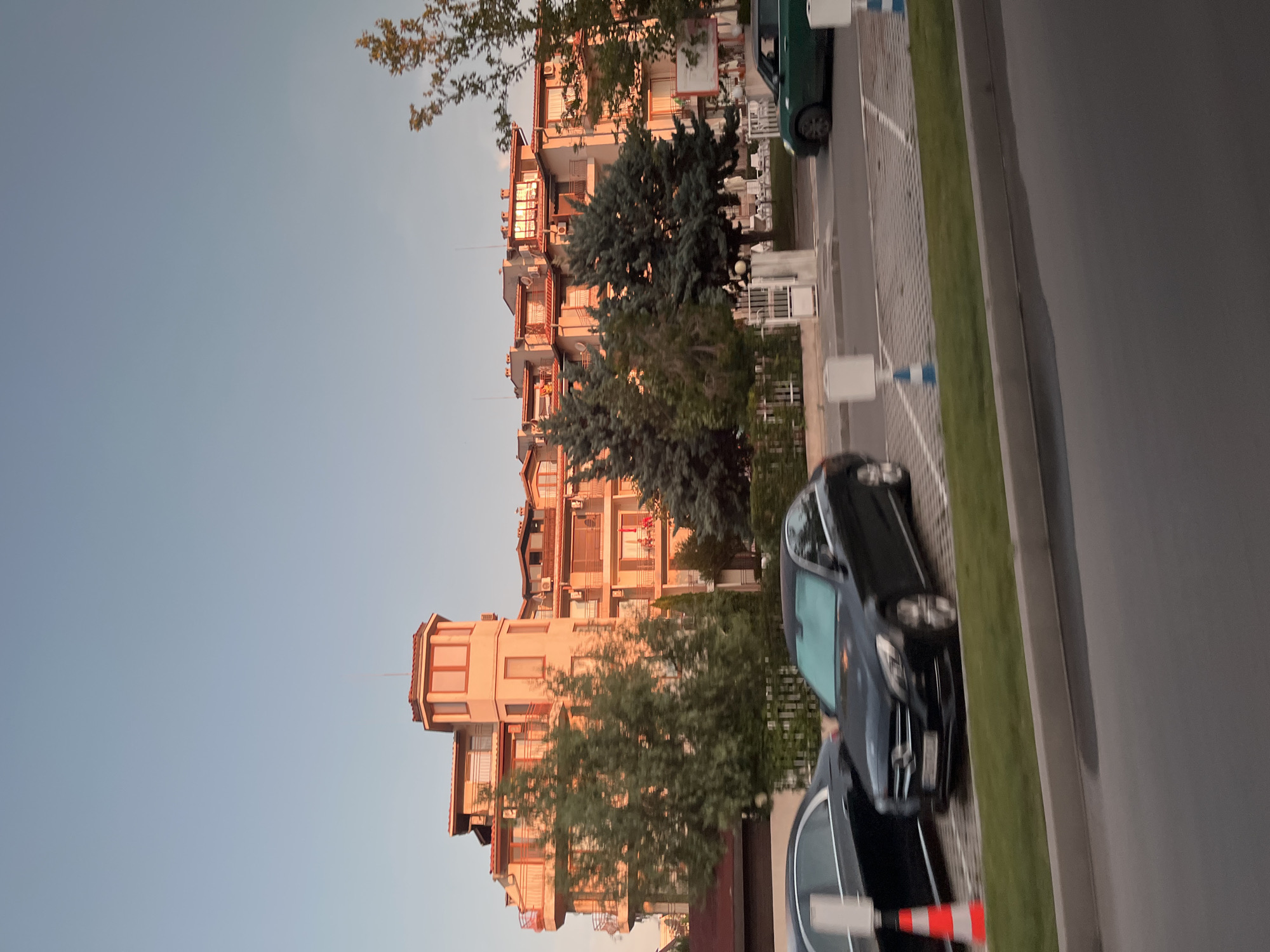 The image shows a street scene in Nesebar, Bulgaria, with a multi-story building in the background and several cars in the foreground. The building, bathed in the warm light of what appears to be sunset, features a distinctive octagonal tower with multiple windows. It has several balconies and is surrounded by trees. A black Mercedes-Benz sedan is parked in a designated parking spot, and another black car is partially visible to its left. A small green car is driving in the background. The street is paved, and there are white and blue bollards marking parking spaces. The sky is a clear, pale blue.