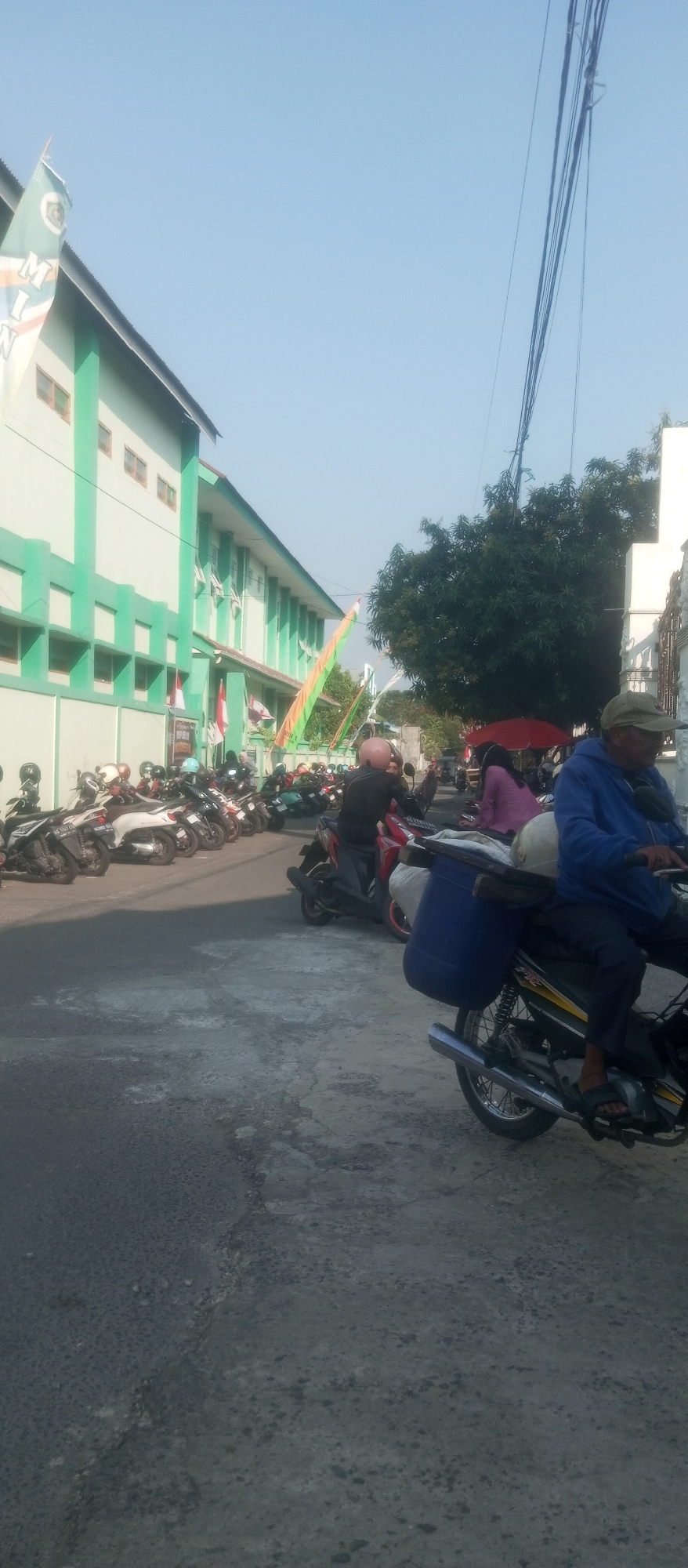The image depicts a street scene in Kediri City, Indonesia, in front of a green and white building. Several motorcycles are parked along the side of the road, and a few people are visible riding or standing near them. One man in a blue jacket and a green cap is riding a motorcycle in the foreground, carrying a blue container on the back. The building appears to be a school or public institution, with windows and a flag pole flying a green and white banner. There are also trees and power lines visible in the background, suggesting an urban or semi-urban environment. The ground is paved with asphalt, and the sky is clear and blue.