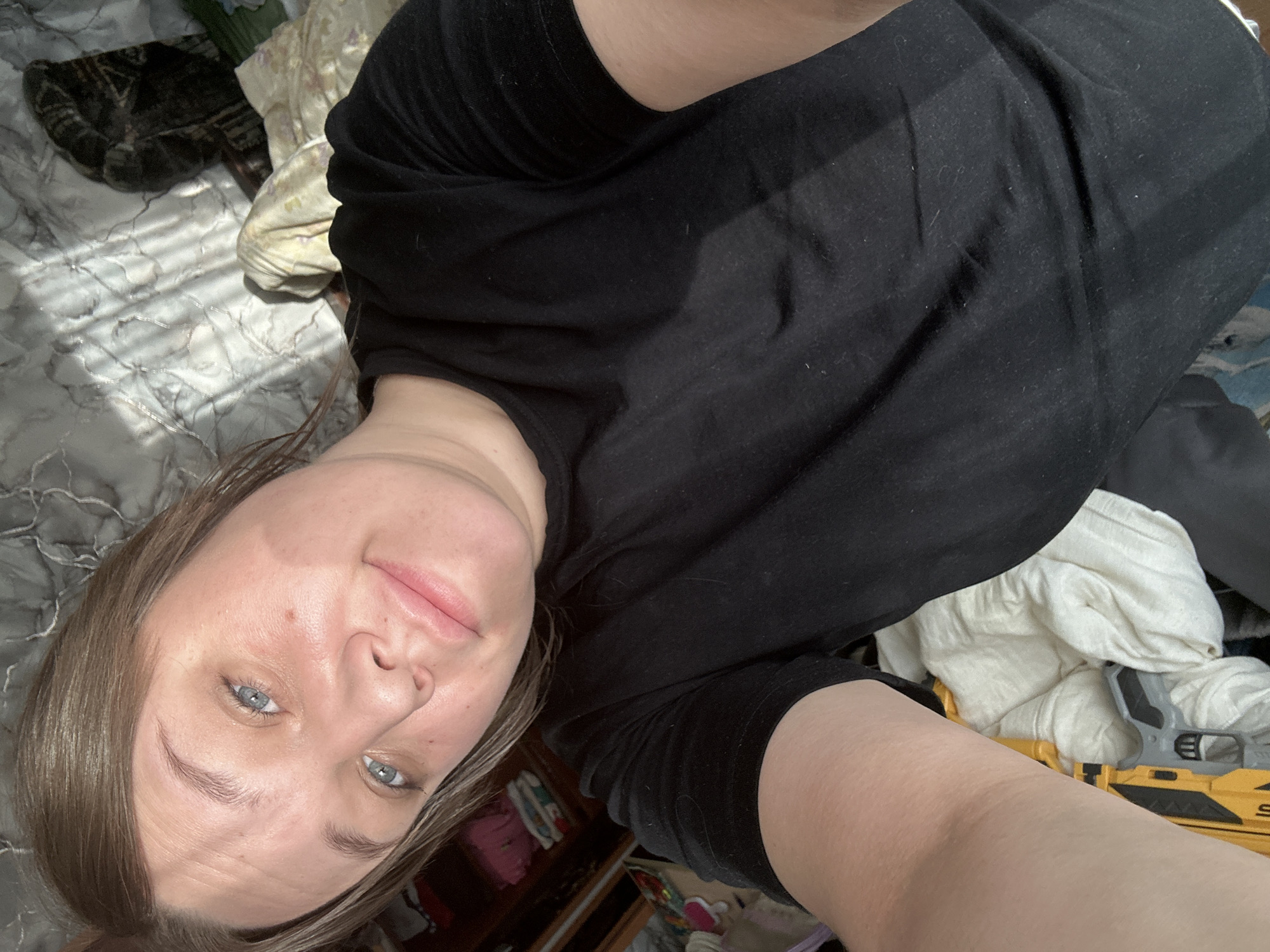A selfie of a woman with light brown hair and blue eyes, wearing a black shirt, is taken indoors. The background includes a patterned blanket, a dark-colored object resembling a cat or cushion, and scattered items including white fabric, part of a yellow and gray toy (possibly a tool), and other personal items, suggesting a cluttered room. The lighting is bright, with some shadows indicating sunlight filtering through blinds. There is no verifiable information to confirm the location as Ольховка, Russia, based solely on the image.