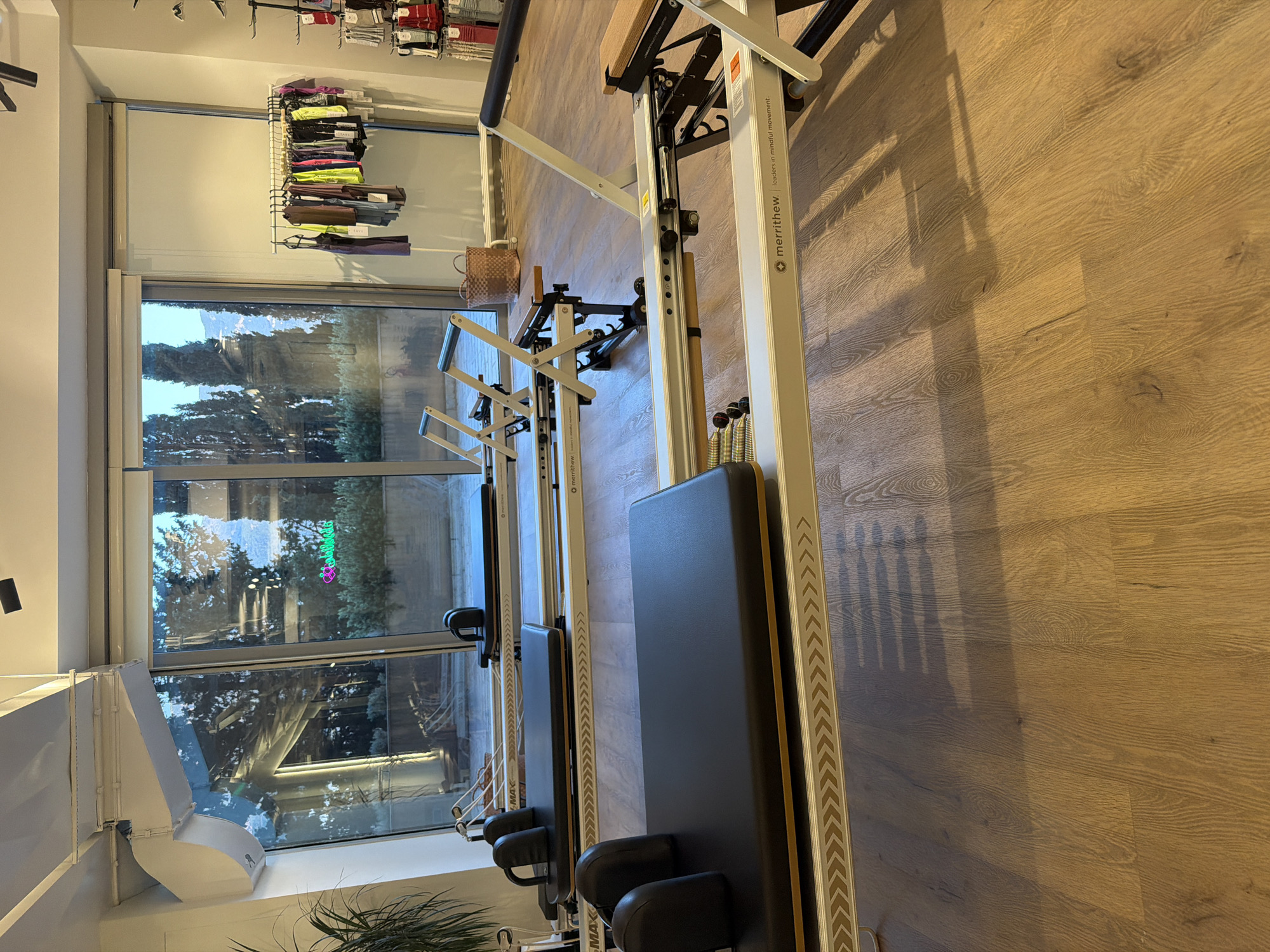 The image depicts the interior of a pilates studio in Boreti, Montenegro. Two Merrithew reformers are the primary focus, positioned parallel to each other on a light brown wooden floor. The reformers are white with black padded carriage and headrests, and wooden accents. The studio features a large window with a view of trees and mountains, suggesting an outdoor environment. To the right of the reformers, a rack displays a variety of athletic wear, and a wicker basket is visible in the background. The lighting is natural, entering from the window, casting shadows on the floor. The overall scene is clean and well-organized, indicative of a professional fitness establishment.