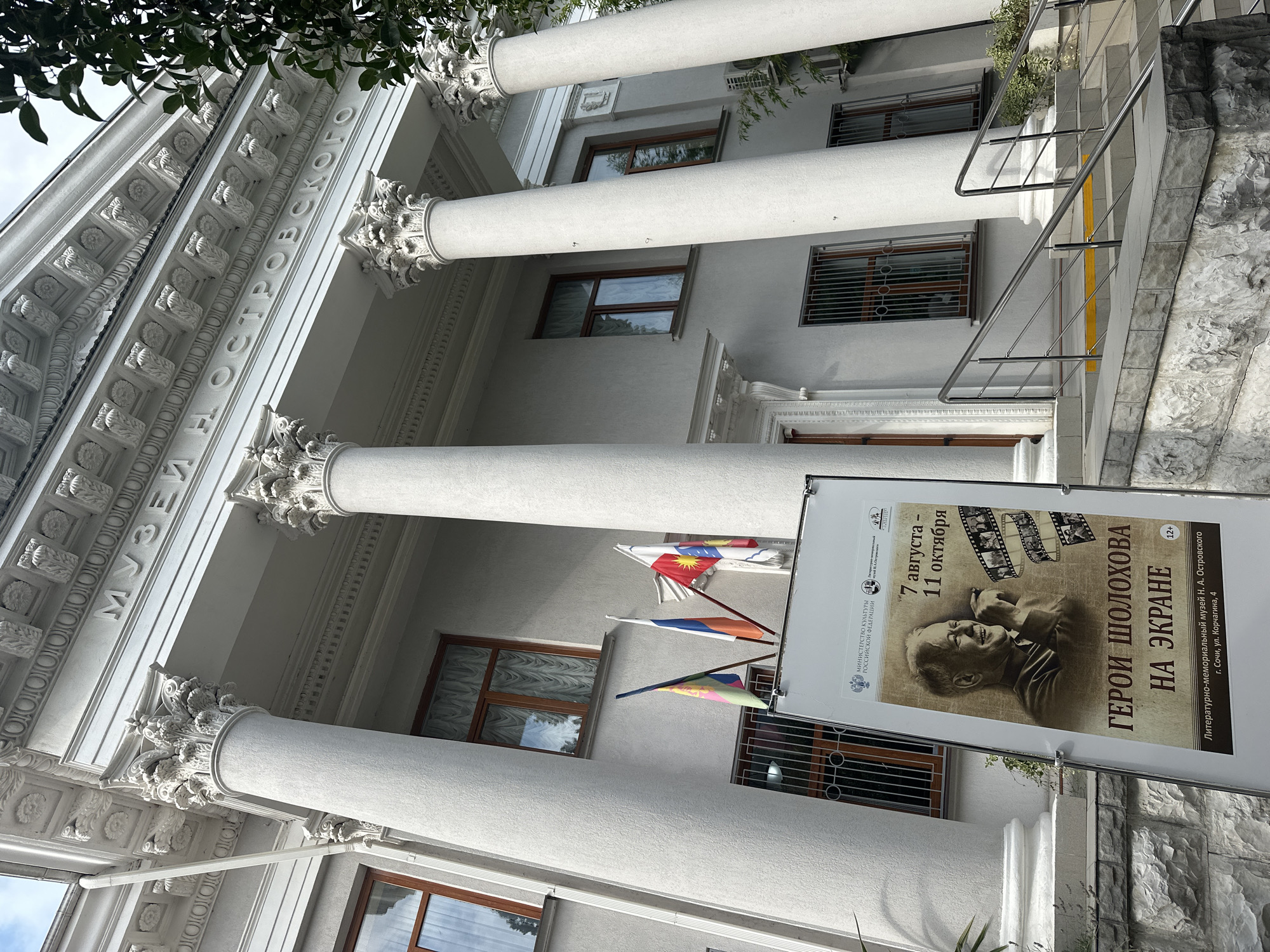 The image depicts the entrance of the N. A. Ostrovsky Literary-Memorial Museum in Sochi, Russia. The building features a grand facade with tall, fluted columns topped with ornate capitals. Above the columns, inscribed in capital letters, is the text "МУЗЕЙ ОСТРОВСКОГО" (Museum of Ostrovsky).
In front of the building, on the steps, is a large white display board advertising an exhibition. The exhibition's title is "ГЕРОИ ШОЛОХОВА НА ЭКРАНЕ" (Heroes of Sholokhov on Screen) and it runs from August 7th to October 11th. A portrait of a man, presumably Mikhail Sholokhov, is featured prominently on the poster, along with film strips. Below the exhibition details, the text specifies the museum's location: "Литературно-мемориальный музей Н. А. Островского г. Сочи, ул. Корчагина, 4" (N. A. Ostrovsky Literary-Memorial Museum, Sochi, Korchagina St., 4).
Three flags are visible flying near the entrance: one with a multi-colored pattern, the Russian flag, and another flag with a red and white design featuring a sun-like emblem. The environment appears to be outdoors, with trees visible on the right side of the frame. The sky is overcast. The architectural style suggests a classical influence. The overall scene is that of a cultural institution open to the public.
