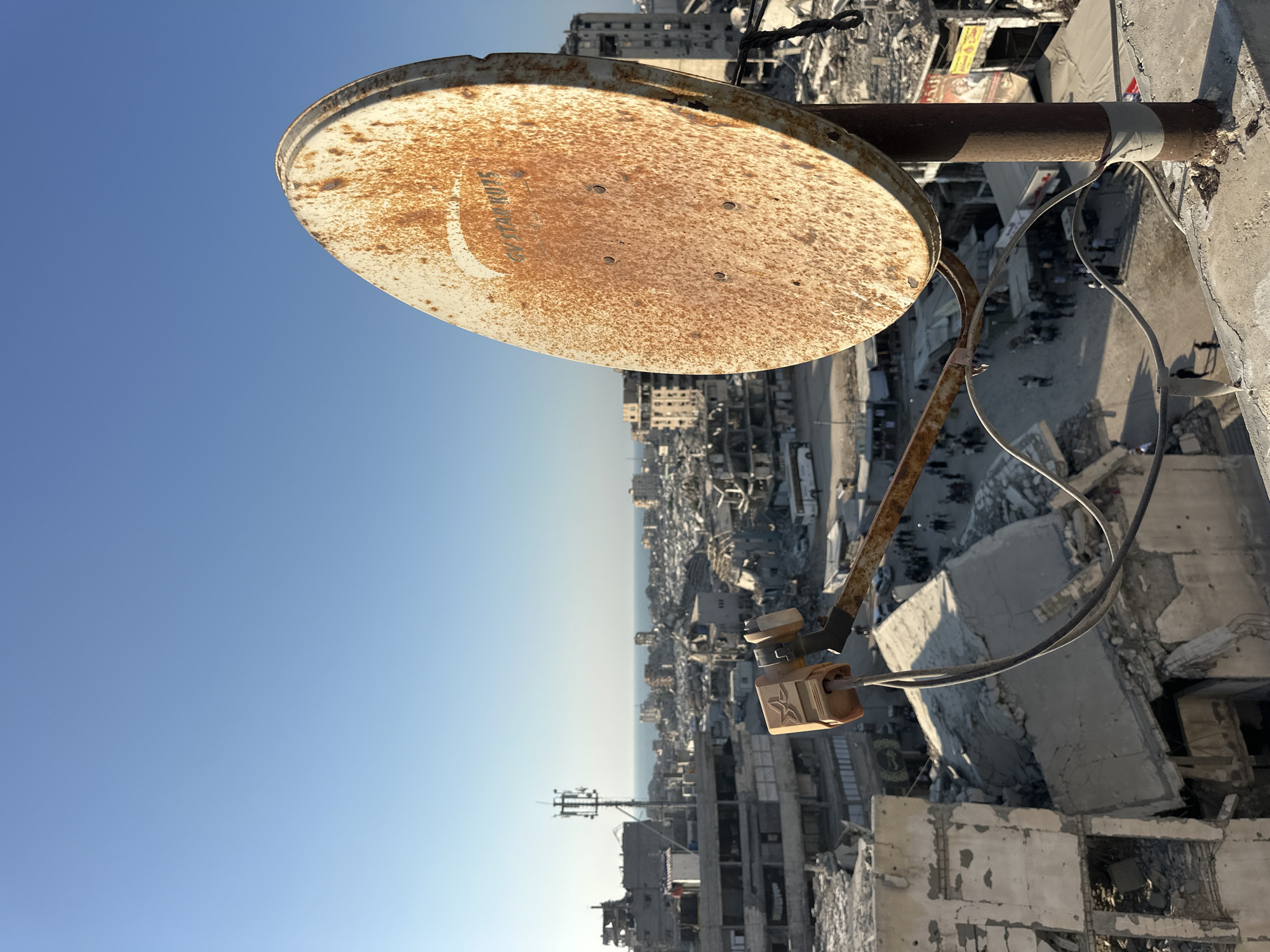 A rusty satellite dish is mounted on a pole atop a concrete structure, overlooking a city in Gaza. The dish is positioned in the foreground, with its circular face bearing signs of wear and tear. In the background, a sprawling urban landscape extends towards the horizon, characterized by buildings in various states of disrepair. Some structures appear to be partially or fully destroyed, with debris scattered across the area.
The city is densely populated, with many buildings closely packed together. In the middle ground, a street can be seen with several people walking and standing around. A few vehicles, including a bus, are also present on the street. The overall impression of the scene is one of a city that has experienced significant damage. The sky above is clear and blue, contrasting with the somber atmosphere of the urban environment. The context of Gaza, Palestinian Territories, implies a region often affected by conflict and destruction, which is evident in the state of the buildings and infrastructure.