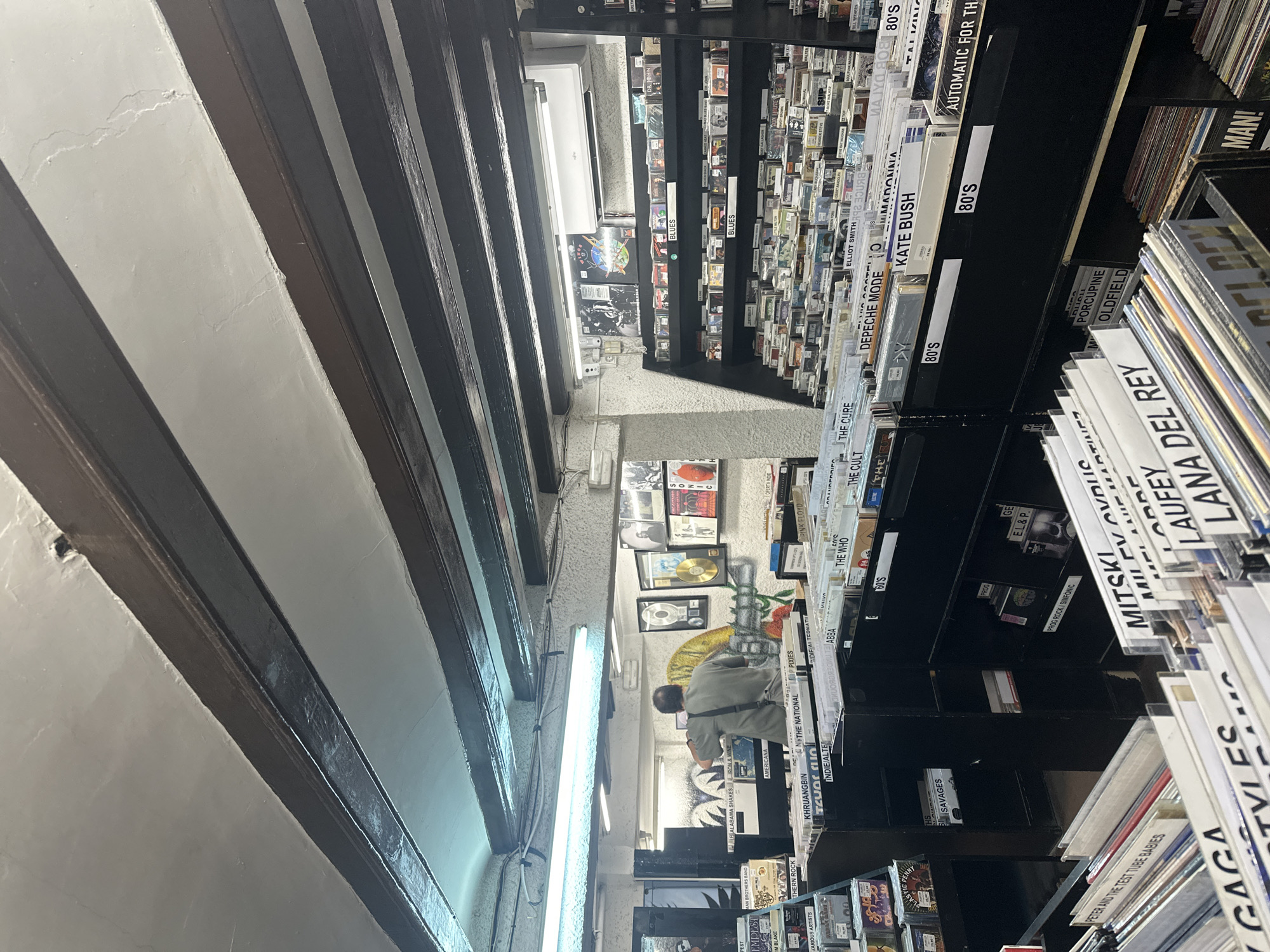 The image shows the interior of a record store, likely in Barcelona, Spain due to the architectural style and dense arrangement of goods. Shelves packed with records stretch into the distance, categorized by genre (e.g., "80's," "Indie/Alter," "Blues") and artist. A man is seen browsing records on the shelves towards the rear of the store. Fluorescent lights illuminate the aisles. The ceiling features a design of alternating dark beams and white plaster, characteristic of some older buildings. The shelves are densely stocked, creating a confined yet immersive environment for music enthusiasts.
