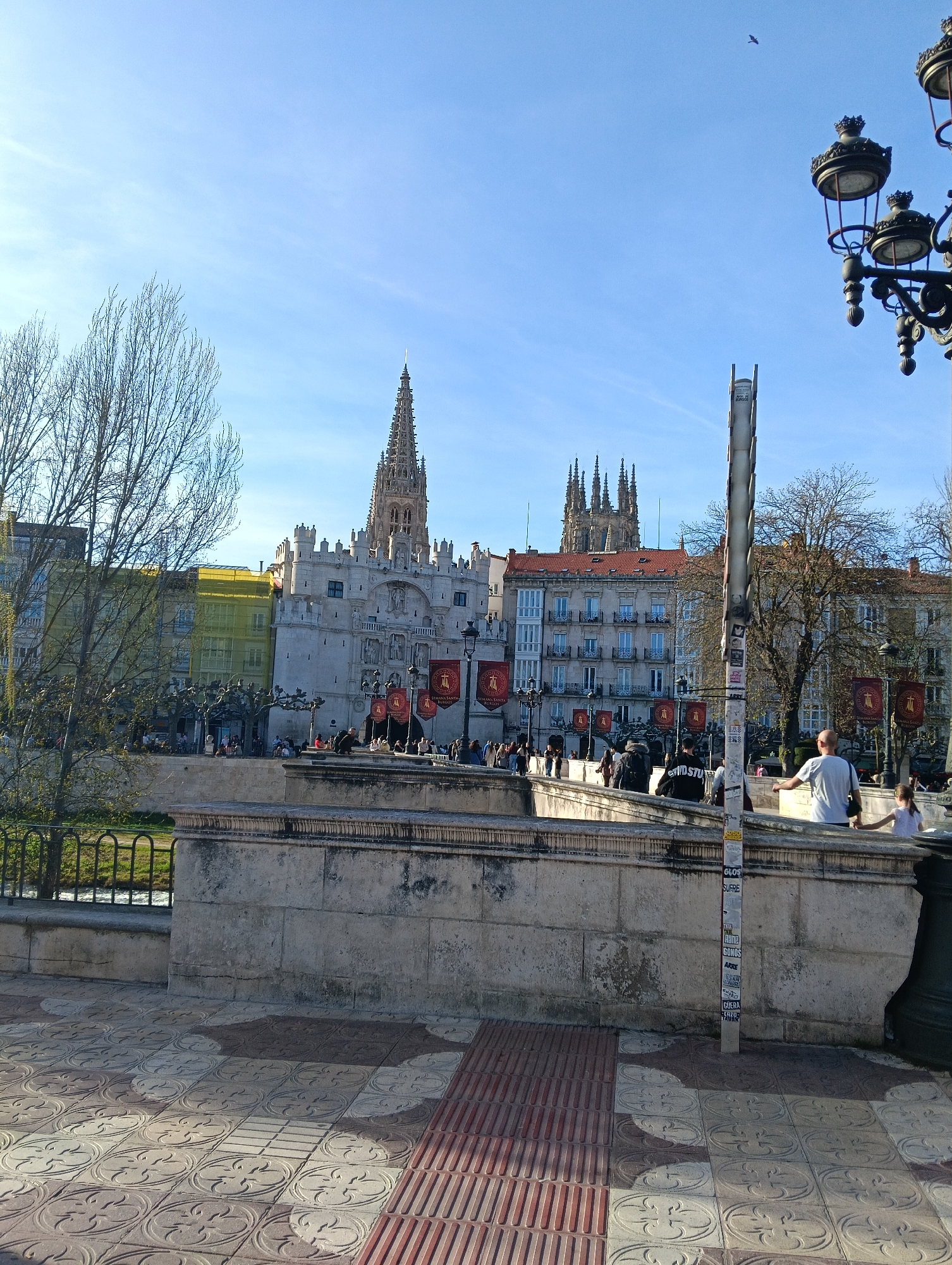 The image captures a daytime scene in Burgos, Spain, featuring prominent architectural landmarks and a lively pedestrian environment.
In the background, the spires of the Burgos Cathedral are visible, along with other historic buildings, some of which appear to be undergoing renovations indicated by scaffolding covered with yellow mesh. Numerous red banners with heraldic emblems are displayed on poles in front of the buildings.
A stone bridge spans the foreground, with people walking across it. Pedestrians of various ages and attire are present, suggesting a public gathering or sightseeing activity.
The pavement in the immediate foreground features decorative tiles and tactile paving with red stripes, likely intended for accessibility. A light post adorned with stickers, along with a portion of an ornate lamppost on the right edge of the frame, add to the urban context. A bird is flying in the clear blue sky.