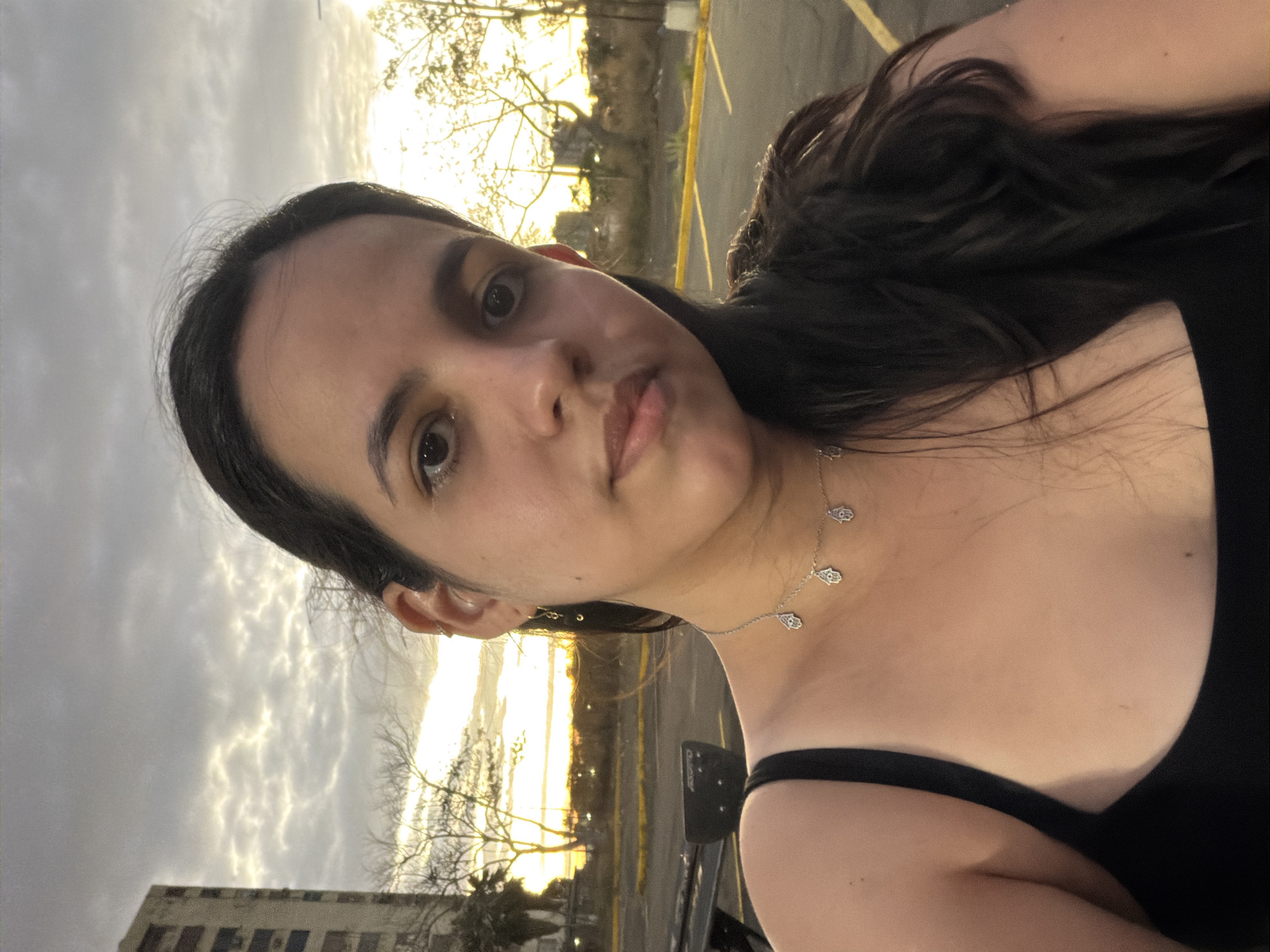A woman is posing for a photograph outdoors. She has long, dark hair and is wearing a black top and a silver necklace with hand-shaped pendants. The background shows a cityscape, including a building, trees, and a cloudy sky illuminated by the setting sun. The location is likely Ciudad Guayana, Venezuela.
