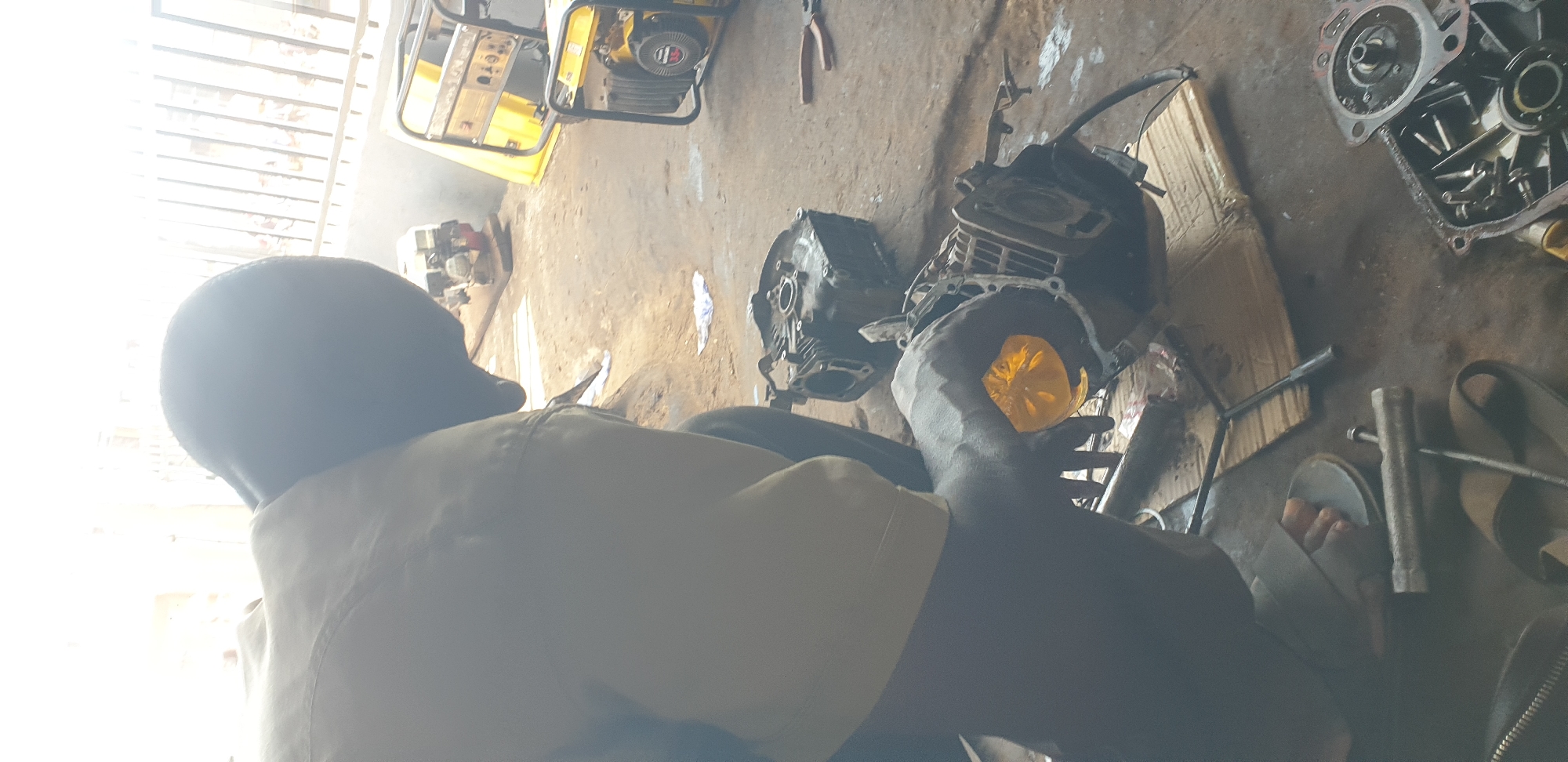A man is kneeling on a dirt floor in what appears to be an outdoor workshop or repair area in Sabon Tasha, Nigeria. He is wearing a tan work shirt and dark pants, and he is working on a disassembled engine. His left hand, holding a yellow object, is placed on the engine block. In the background, there are several generators, one yellow and black, and another partially visible behind it. Tools such as pliers and a T-handle wrench are scattered on the ground around the engine parts. The scene is lit by bright, natural light, creating a harsh glare in the upper portion of the image.
