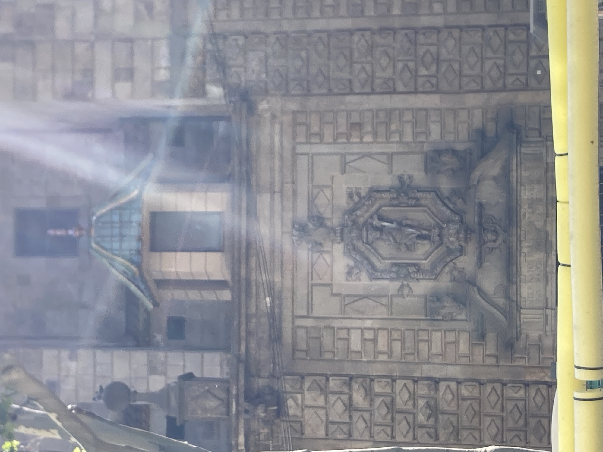 This media file shows a close-up view of an ornate building facade, likely in Barcelona, Spain. The image is dominated by intricate stone carvings and architectural details. A central element is a large, oval-shaped relief depicting a bird in flight within a decorative frame. The surrounding stonework features a repeating pattern of squares and diamonds, with horizontal bands of rectangular shapes.
The lighting suggests it is daytime, with sunlight casting some glare and lens flare onto the scene, particularly on the left side. This glare obscures some of the finer details of the building's upper sections. On the right side of the image, two large, bright yellow cylindrical objects, possibly scaffolding poles or construction materials, are prominent. Their presence indicates that some form of construction or renovation might be underway.
On the far left, a section of a building with windows and a blue-tinted, angled structure can be seen. This part of the structure has a more modern appearance compared to the ornate stonework. The overall impression is of a building with a rich historical architectural style, possibly a cathedral, a palace, or a historic government building, juxtaposed with modern elements or temporary structures.
A faint inscription is visible on the stonework on the right side of the image, behind the yellow poles, but it is too blurry to be deciphered. The image is captured from a slightly elevated perspective, looking up towards the facade. The scene is static, with no people or overt activity visible, focusing entirely on the architectural elements of the building. The weather appears to be clear and sunny.