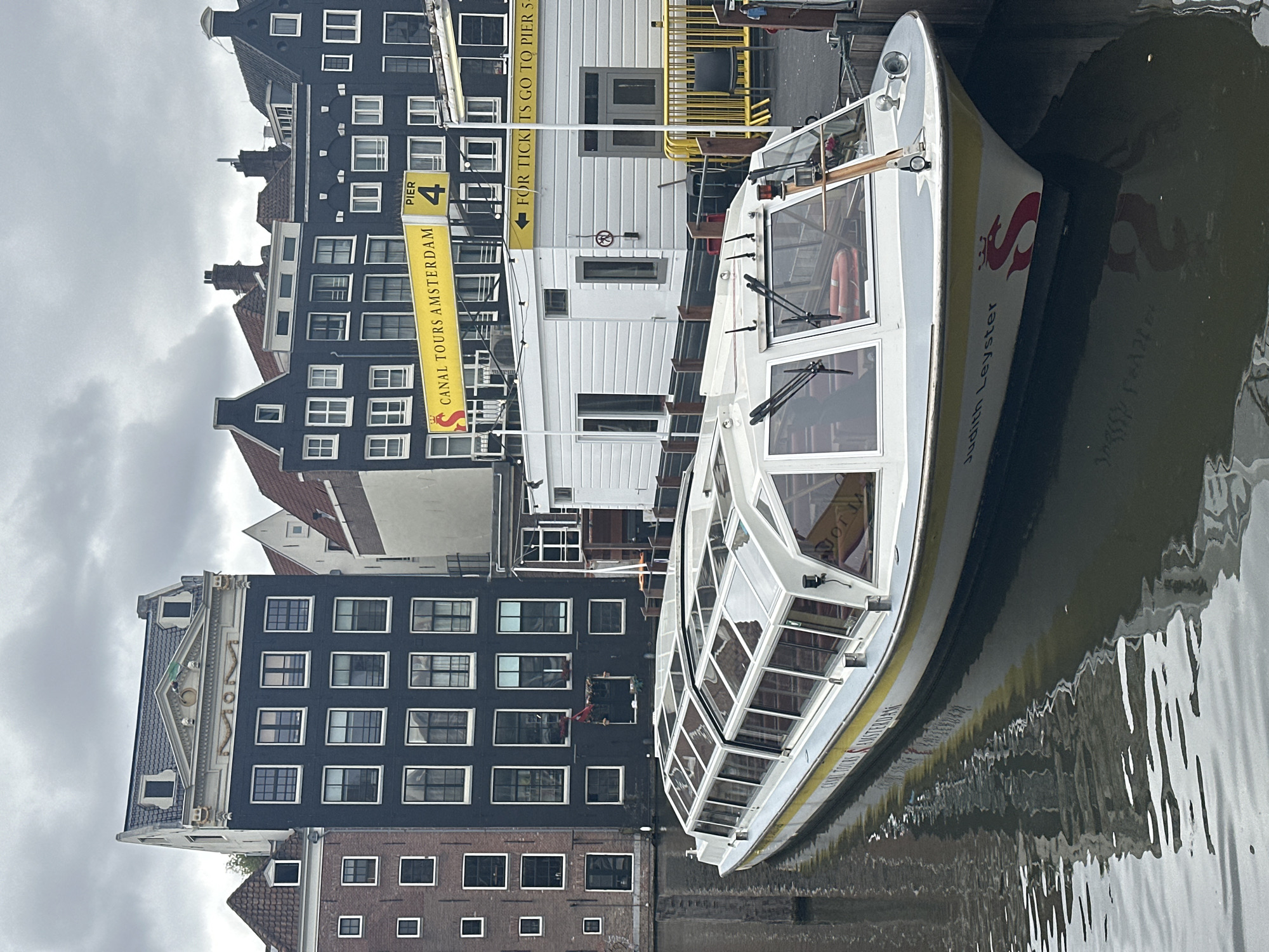 The image shows a canal boat named "Judith Leyster" docked in a canal in Amsterdam, Netherlands. The boat has a glass roof and sides, and is painted white with a yellow and grey stripe. It is surrounded by traditional Dutch canal houses, characterized by their narrow facades and steep roofs. A yellow sign advertising "CANAL TOURS AMSTERDAM" and "PIER 4" is visible in the background, along with a smaller sign indicating "FOR TICKETS GO TO PIER 5". The sky is overcast, contributing to a muted color palette of the scene. The water in the canal reflects the buildings and the boat, creating a shimmering effect. There are no people clearly visible in the frame.