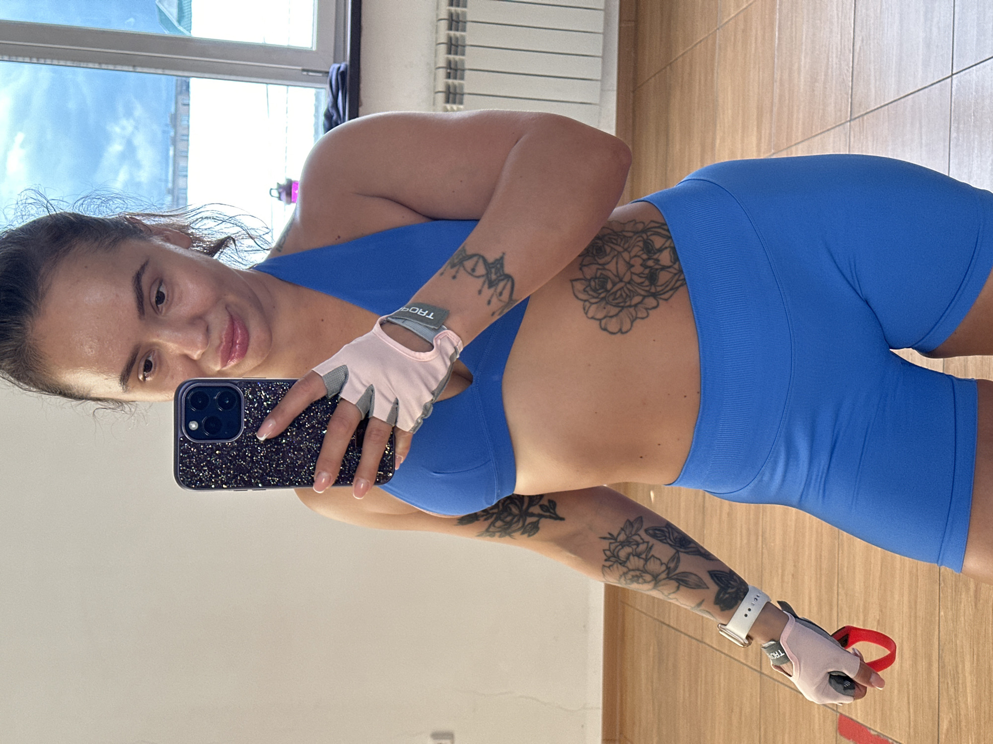 A woman is taking a mirror selfie in what appears to be a fitness studio or gym in Yuzhnouralsk, Russia. She is wearing a blue sports bra and blue workout shorts. She has several tattoos on her arms and torso, including floral designs and what looks like a lion's head. On her left wrist, she is wearing a white smartwatch, and on her hands, she has light pink fitness gloves. One of her hands is holding a red resistance band loop. The background shows a white wall, a wooden floor, a window with a radiator below it, and a glimpse of a sky with clouds outside the window.