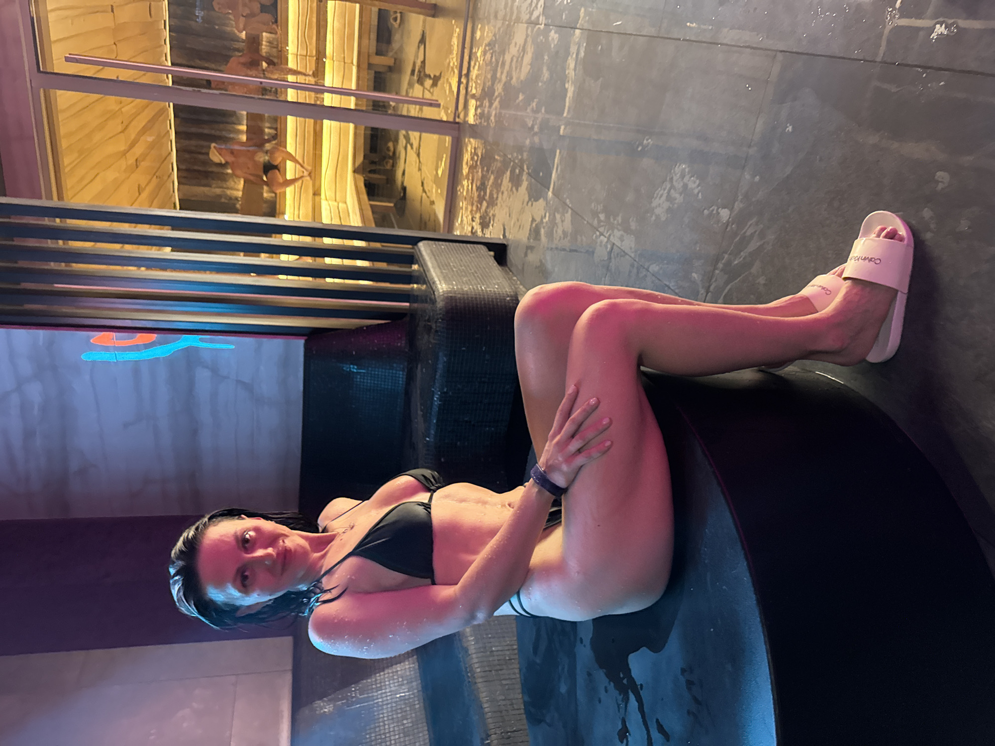 An indoor, vertical image shows a woman reclining on a dark, curved structure in what appears to be a spa or pool area. She is wearing a black bikini and white "Calvin Klein" slides on her feet. Her skin is wet, and water droplets are visible on her body and the surrounding dark, tiled floor.
The woman is positioned with her legs bent and her hands resting on her thighs, looking towards the camera with a slight smile. A dark watch or bracelet is visible on her left wrist, and she wears a delicate necklace.
The setting is characterized by dim, artificial lighting with noticeable pink and blue hues. The floor is wet and reflective. In the background, behind a low dark barrier, there's a glass partition. Through this partition, a reflection reveals wooden paneling, strongly suggesting a sauna or steam room. Figures of other people are dimly reflected within this wooden-paneled area, appearing to be sitting or standing. The overall atmosphere suggests relaxation and leisure in a water-related facility.