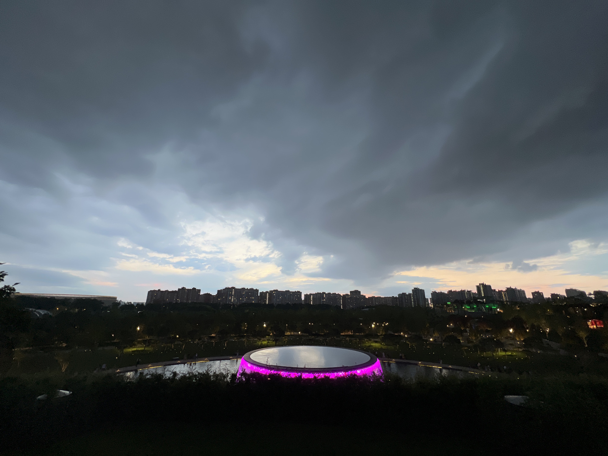 The image captures a twilight scene in Krasnodar, Russia, focusing on a circular, illuminated structure at the foreground, which appears to be a fountain or a stage feature with vibrant pink lighting. Beyond this structure, a park or garden area is visible, with trees and pathways illuminated by scattered lights. A body of water surrounds the central structure. In the background, a city skyline is silhouetted against the darkening sky, featuring numerous apartment buildings and some with green lighting. The sky itself is dramatic, filled with heavy, dark clouds, suggesting an approaching storm or the aftermath of one. The overall impression is a blend of urban landscape and natural elements, with man-made illumination providing points of interest against the dimming natural light and imposing cloud cover.