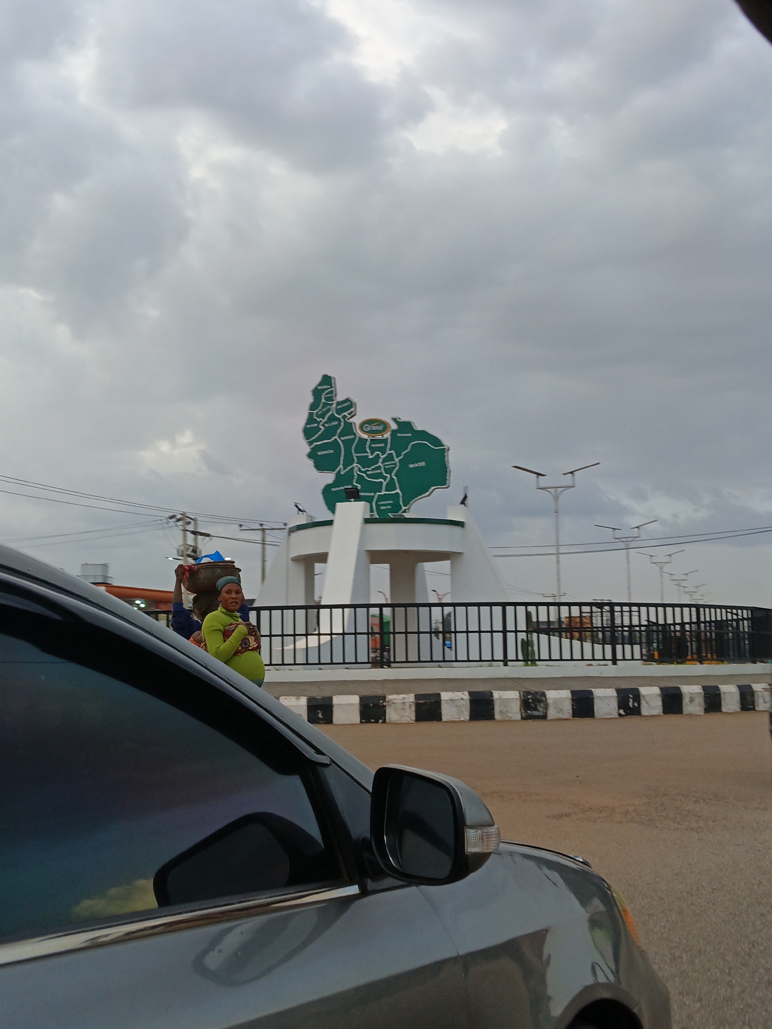 A medium shot captures a street scene in Kuru Dungus, Nigeria, under a cloudy sky. In the background, a large, green monument shaped like a map of a region, possibly a state, is mounted on a white pedestal. The map is segmented and labeled with place names, with "Grand" visible on a small circular emblem. Several streetlights with double arms are visible behind the monument. A black metal fence with vertical bars separates the monument area from the road. In the mid-ground, a woman wearing a green head covering and a bright green sweater is standing and looking towards the camera. She has a determined expression and is holding her arms across her stomach. To her left, another person, partially obscured, appears to be carrying an object on their head. In the foreground, the dark, tinted window and side mirror of a gray car dominate the lower left portion of the frame, reflecting the cloudy sky and some of the surroundings. The road surface in the foreground is paved.