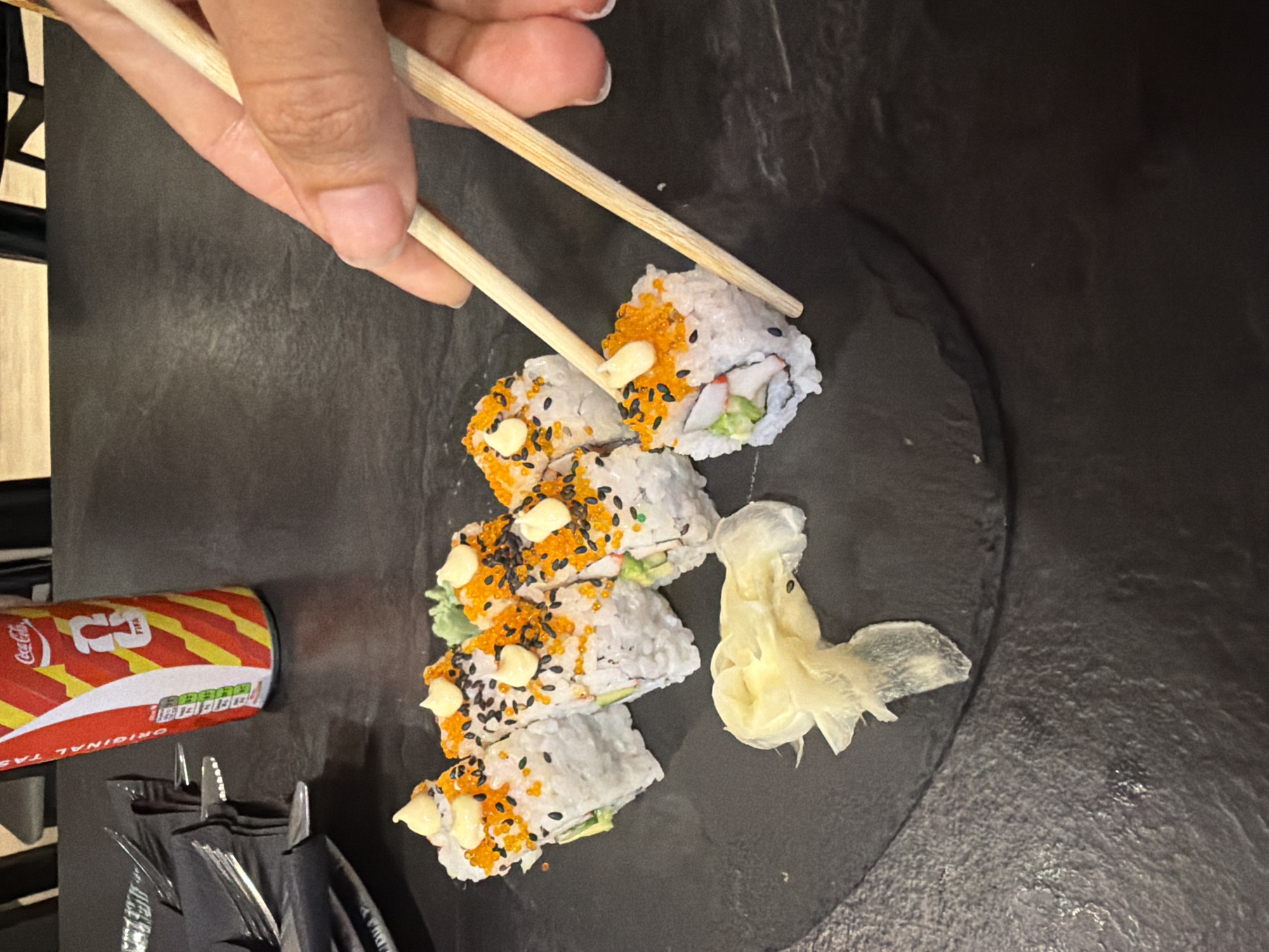 In the city of Kiti, Cyprus, a hand holds chopsticks, picking up a piece of sushi from a black slate plate. The sushi rolls are white and cylindrical, topped with orange roe, black sesame seeds, and dollops of white sauce. A small amount of green wasabi is visible on the plate, as is a piece of pickled ginger. To the left of the sushi, a black napkin is folded around silverware. In the background, a red and white Coca-Cola can with a FIFA logo is standing upright. The scene is set against a dark, textured surface, likely a table.