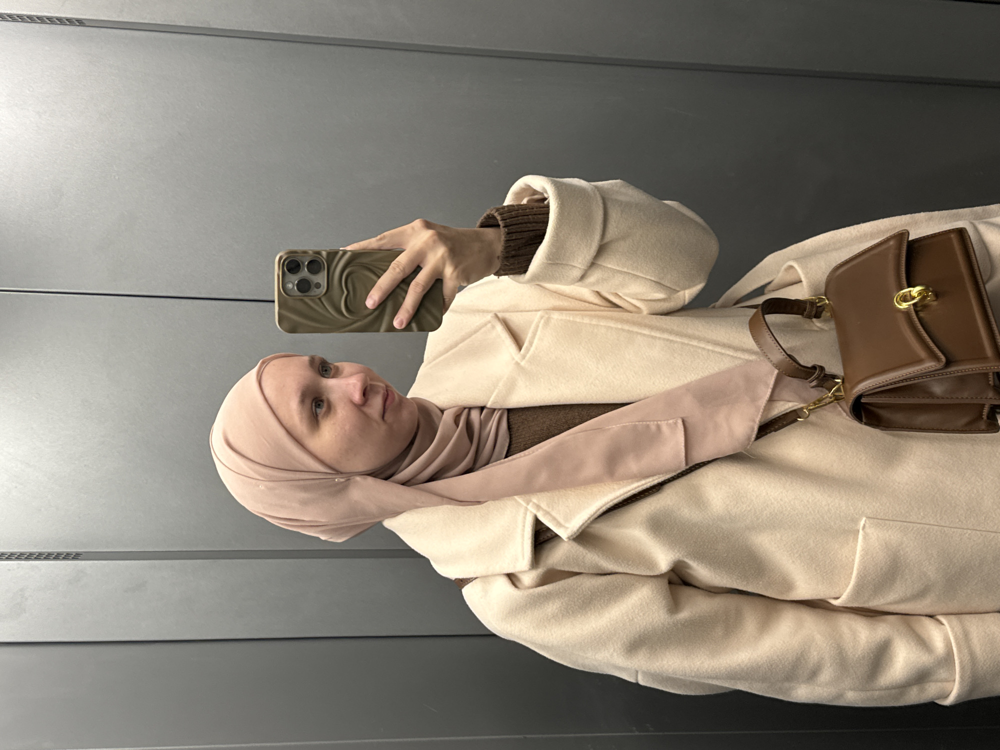 A woman in a hijab is taking a selfie in what appears to be an elevator. The background is gray, indicating the elevator's interior. She is wearing a light-colored coat, a brown sweater, and a matching hijab. She is holding a phone with a decorative case. A brown leather purse is slung over her shoulder. The location in Chaikovsky, Russia is not evident from the image.