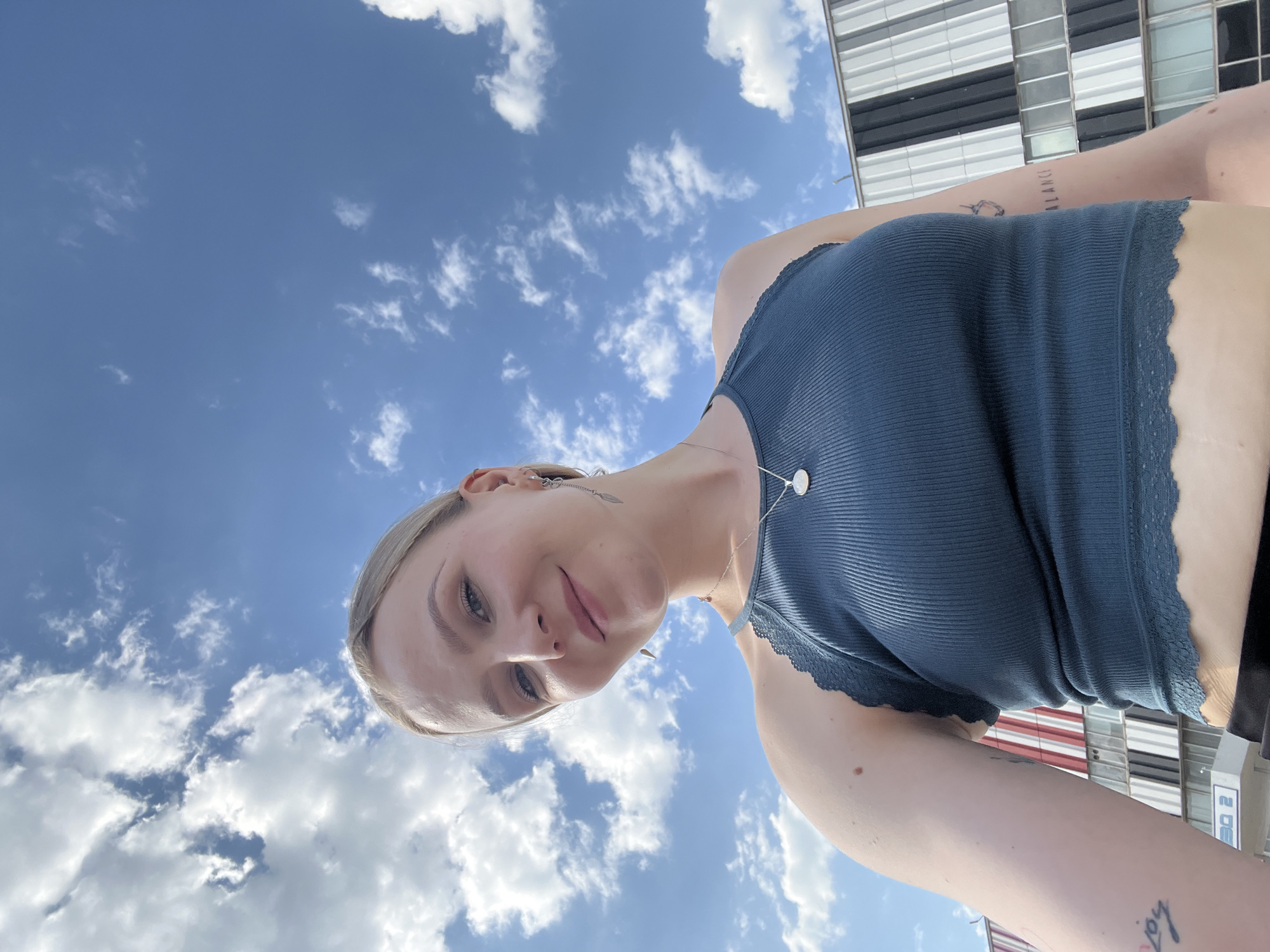A low-angle, tilted, full-body selfie shows a fair-skinned person with blonde hair wearing a dark teal tank top with lace trim, a silver necklace with a circular pendant, and various earrings, against a backdrop of a blue sky with scattered clouds. The person is smiling slightly, looking down at the camera. A building with horizontal white and black lines is visible in the upper right corner. In the person's upper right arm, the word "BALANCE" is tattooed on the person's skin. This image was taken in the city of Khimki, Russia.