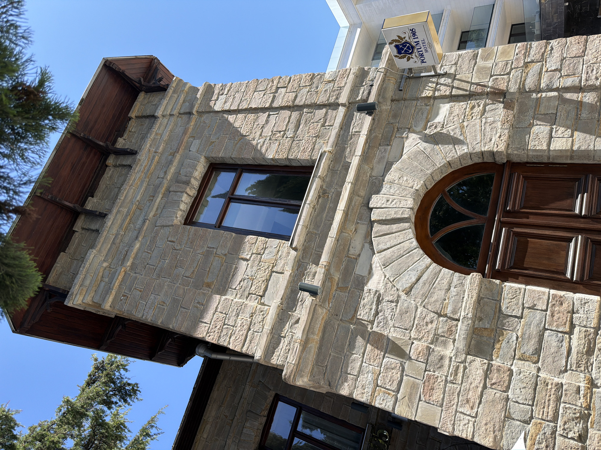 The image captures a stone building in Sochi, Russia, with a clear blue sky overhead. The building features a robust stone facade with visible mortar lines. A large wooden arched doorway is centrally located, framed by rough-hewn stones. Above the doorway, a window with a dark wooden frame reflects the surrounding greenery. A sign reading "PORTUM 1905 HOTEL" is mounted on the right side of the building. To the right of the stone building, a modern white hotel with multiple balconies and glass railings is visible, suggesting a juxtaposition of old and new architecture. Tree branches with green foliage partially obscure the top of the stone structure. The lighting indicates it is daytime, with distinct shadows cast on the building's surface. There are no people visible in the scene.