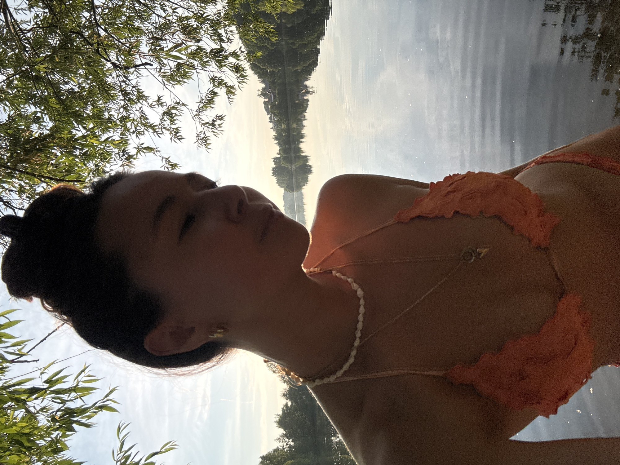 A young woman with her hair in a bun is standing next to a lake during sunset. She is wearing an orange bikini and several necklaces. The lake is surrounded by trees and in the distance there are houses. The sun is setting, casting a warm glow over the scene. The woman is looking away from the camera, towards the lake.
