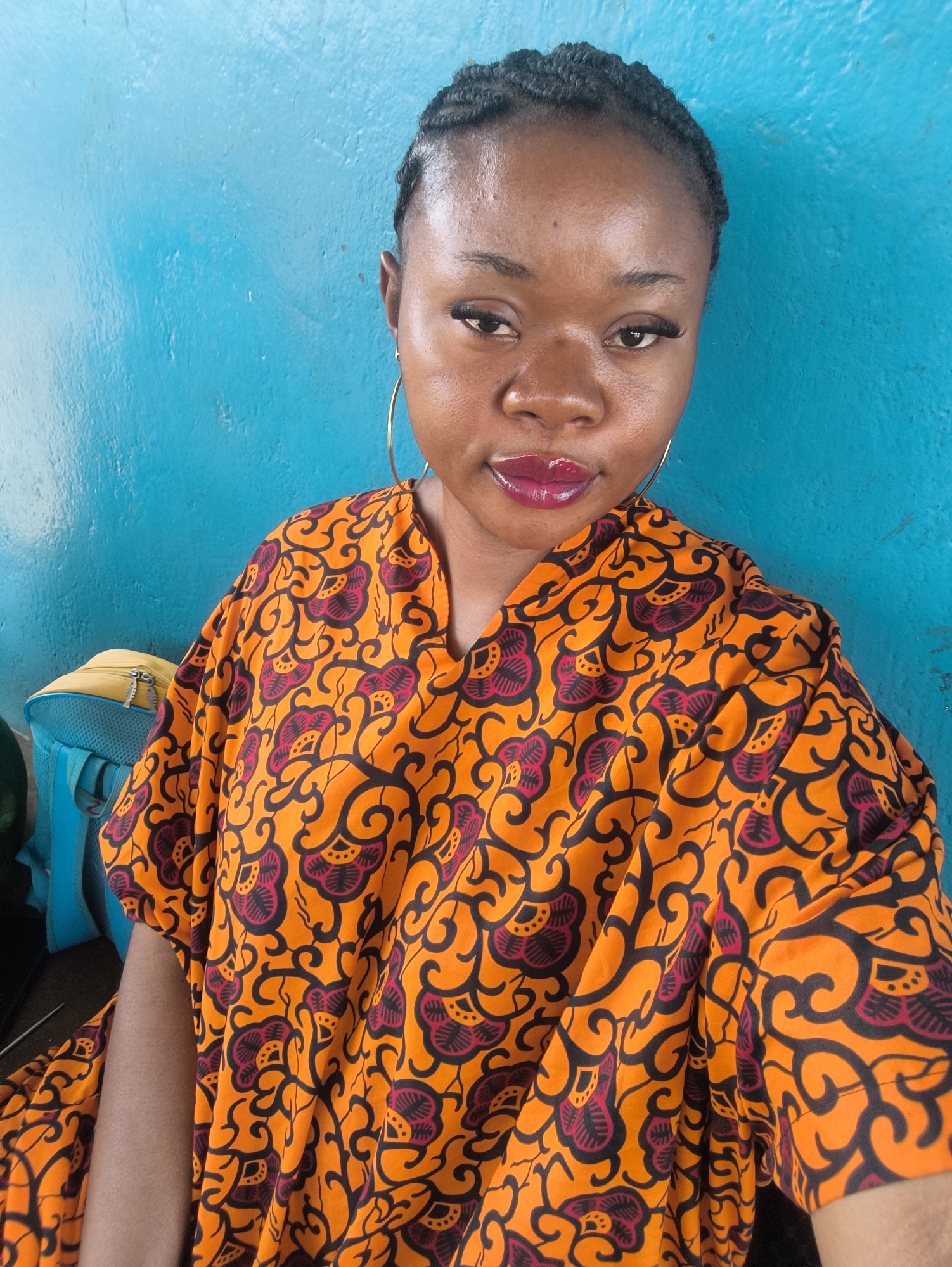 A medium-shot portrait of a young woman with braided hair is set against a light blue wall. She wears large gold hoop earrings and a bright orange patterned dress with black and dark pink floral designs. Her lips are glossy with a deep red lipstick. To the left, partially visible, are a light blue and yellow backpack. The location is likely a home or indoor setting in Jos, Nigeria. The image is taken with a slightly upward angle.