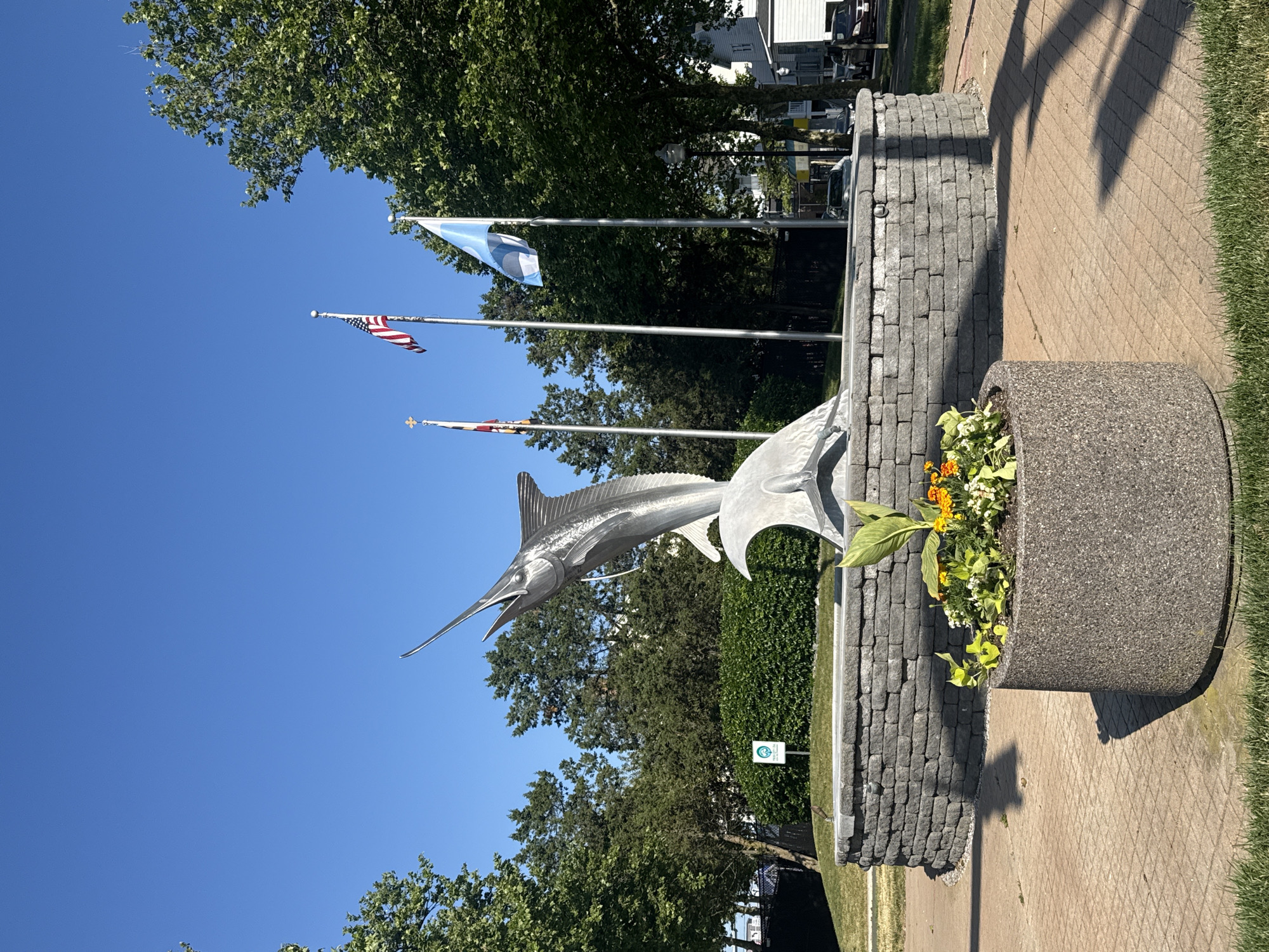The image depicts a monument in Ocean City, Maryland, United States, featuring a silver marlin statue mounted on a gray brick pedestal. In the background, three flags are visible atop poles: a blue and white flag, the American flag, and the Maryland state flag. A flower pot with green and yellow plants sits next to the monument, positioned on a brick pathway with grass on either side. Trees and buildings can be seen in the background under a clear blue sky.